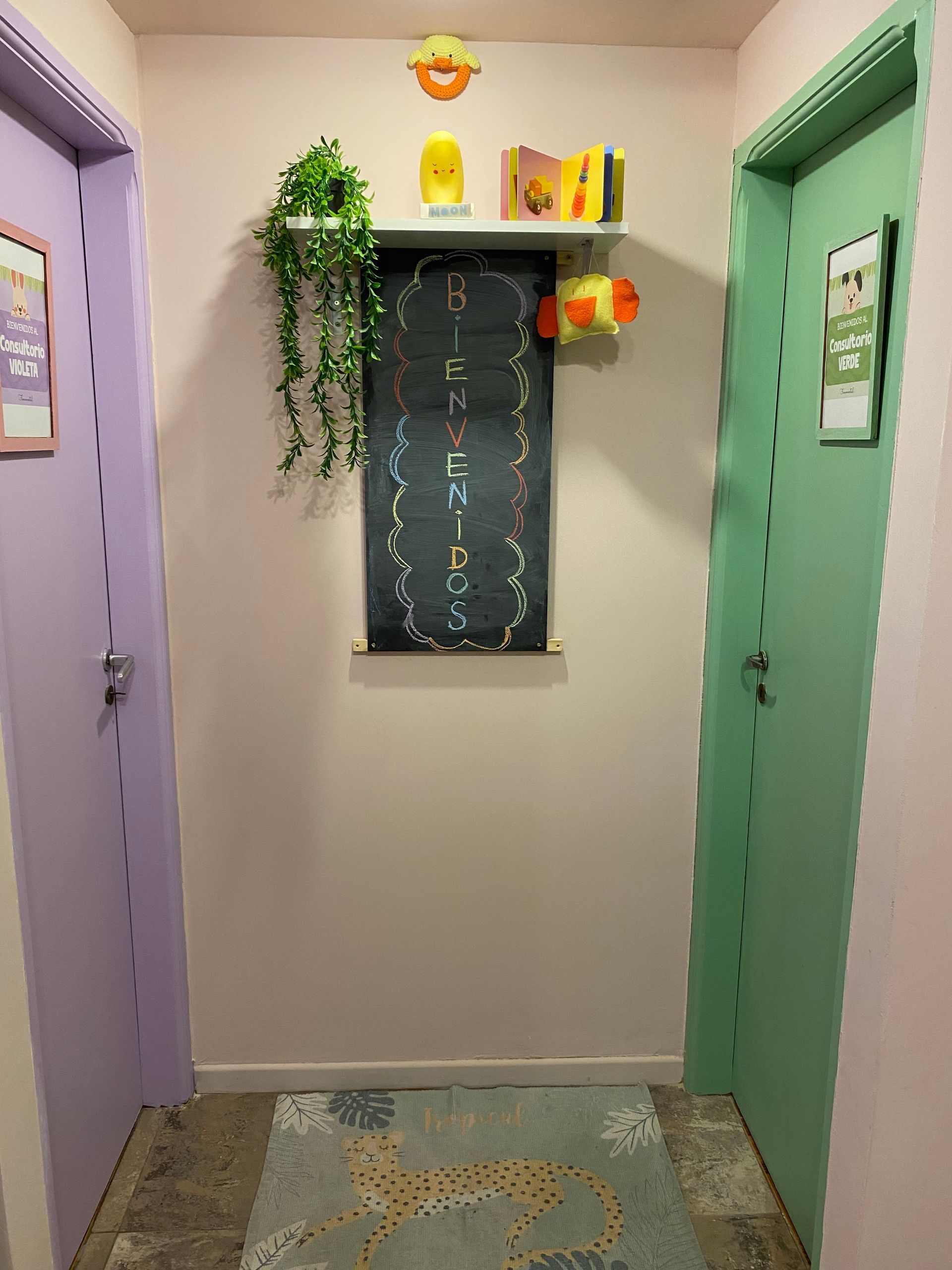 Pasillo con paredes rosas, puertas moradas y verdes, pizarra y estantería decorativa.