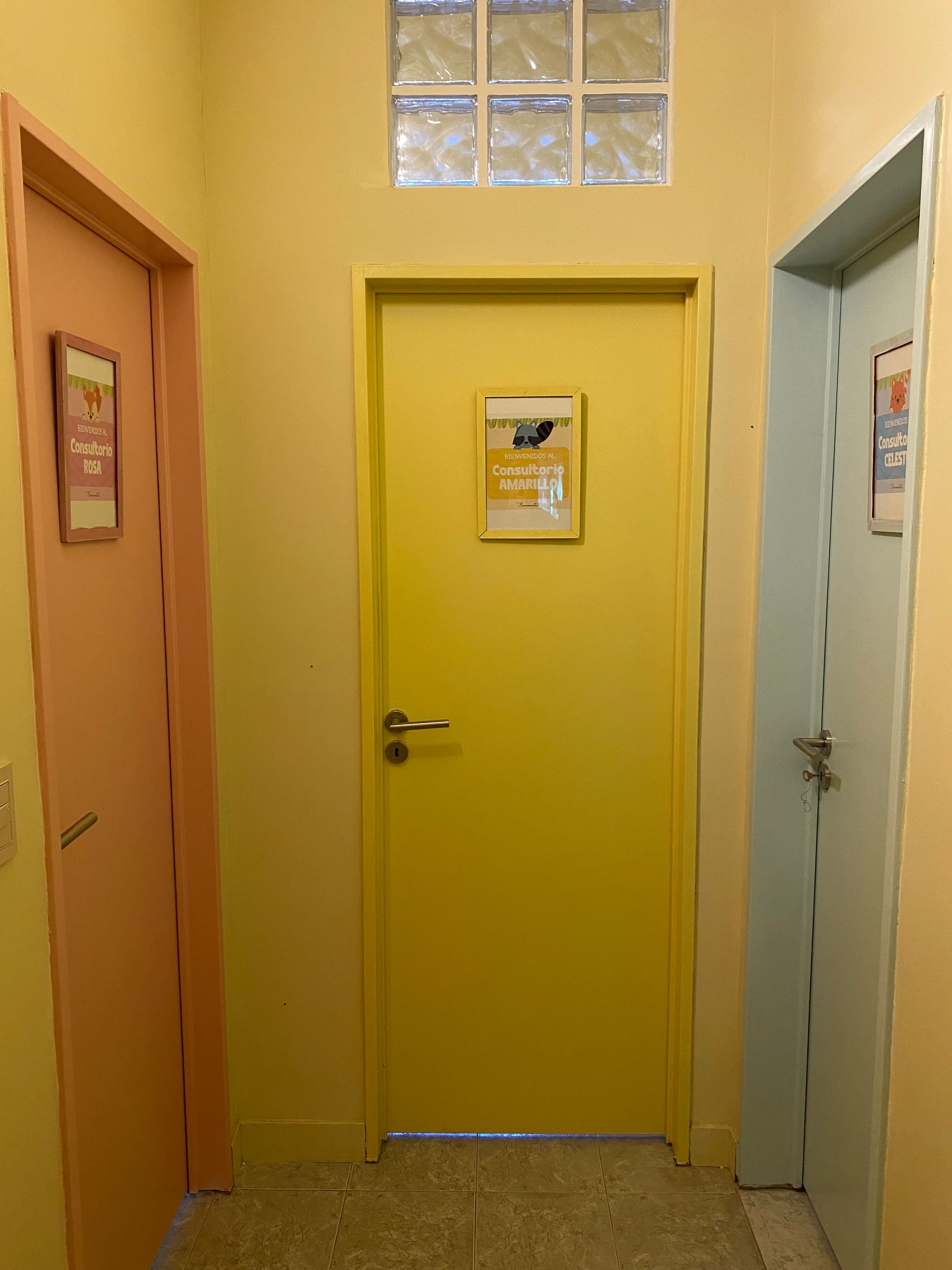 Pasillo con tres puertas de colores, paredes amarillas y una ventana de bloques de vidrio.