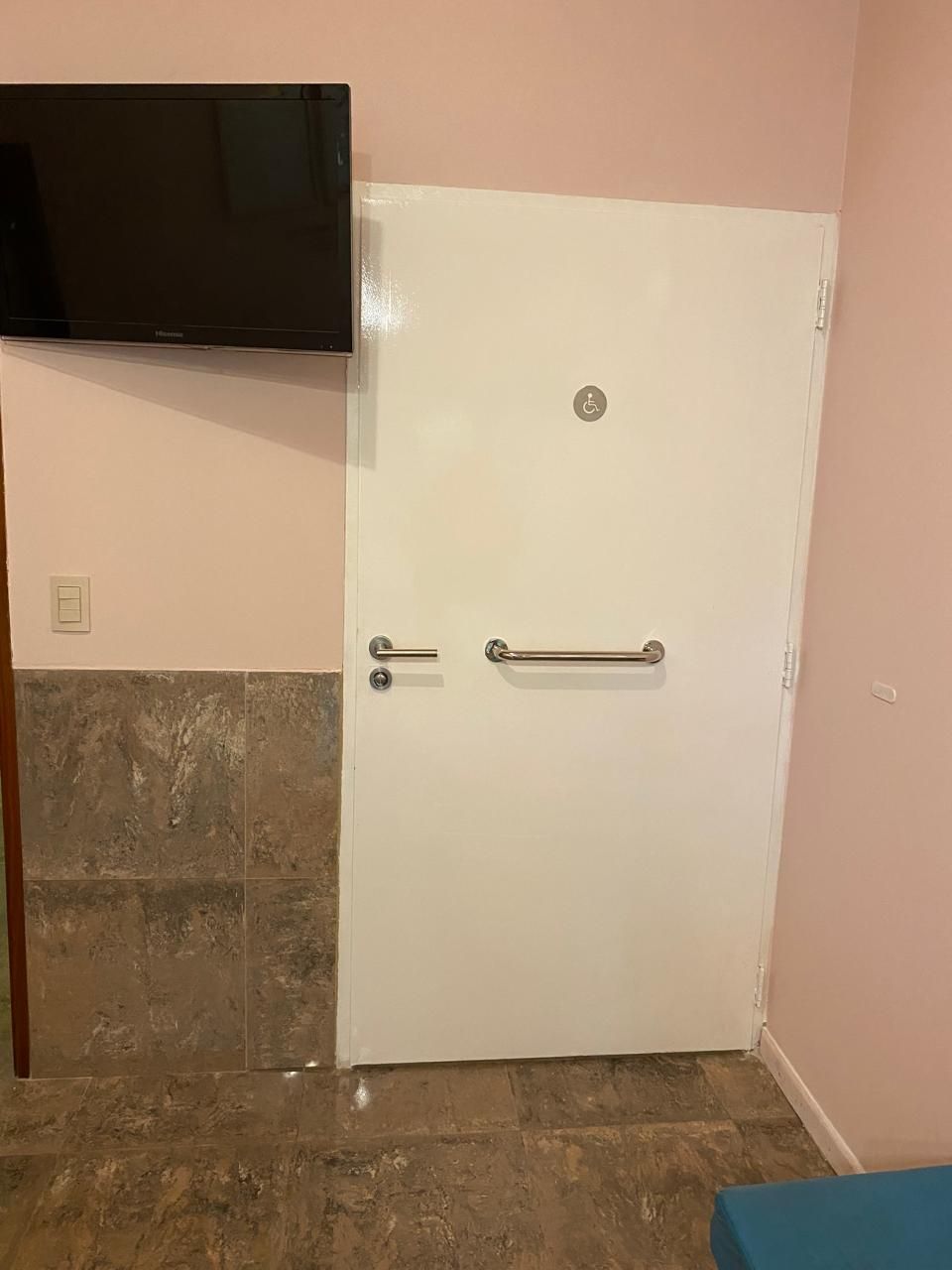 Puerta blanca con barra de apoyo y tirador sobre una pared rosa, cerca de un televisor.