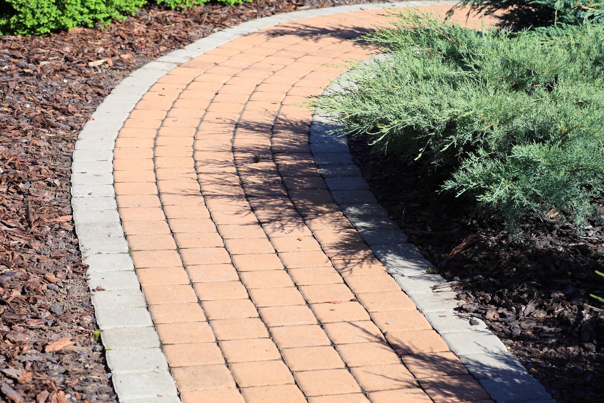 Brick walkway curving through a garden, bordered by gray bricks and mulch.