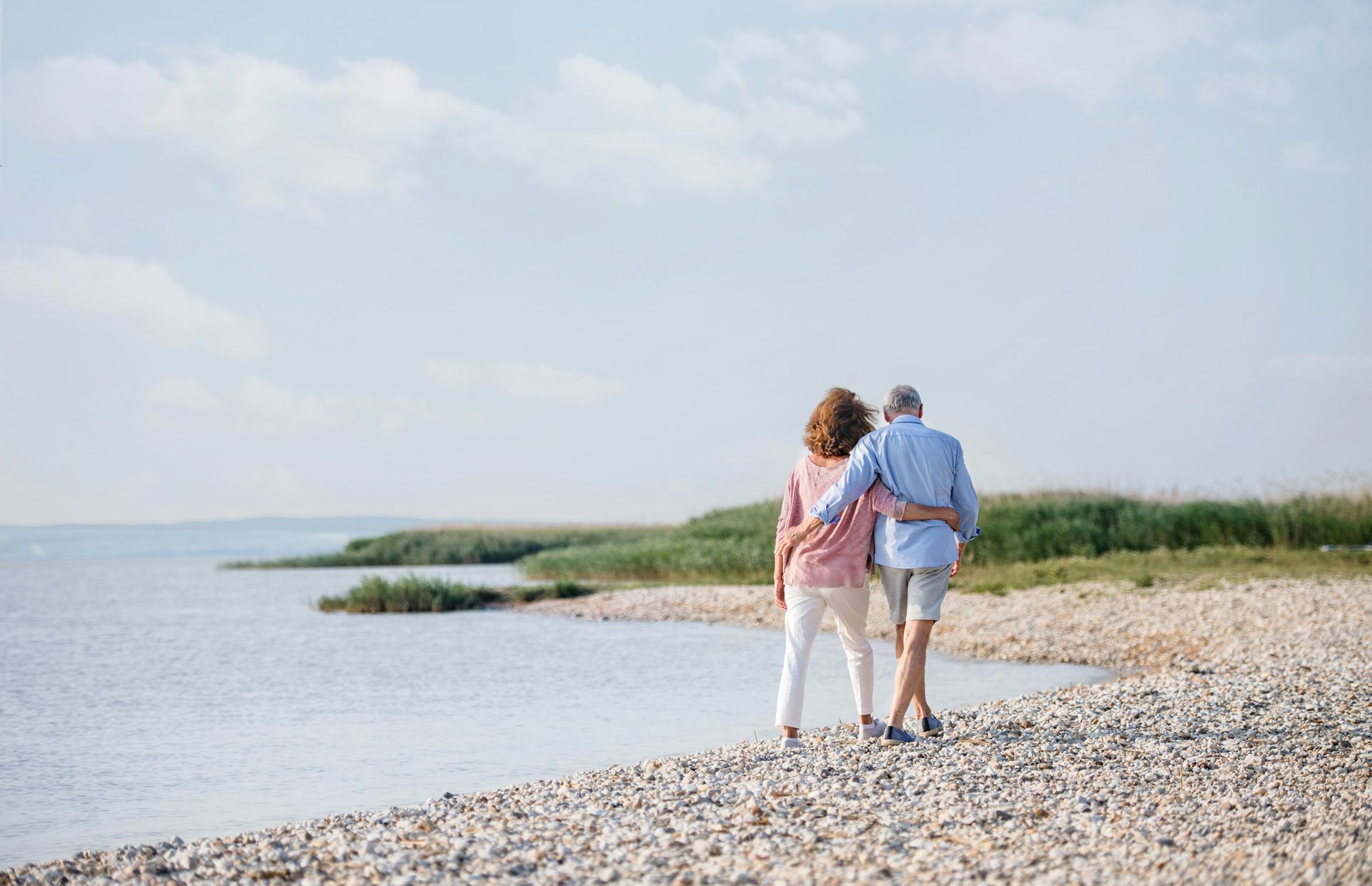 Two people walking along a rocky beach beside calm water under a pale sky