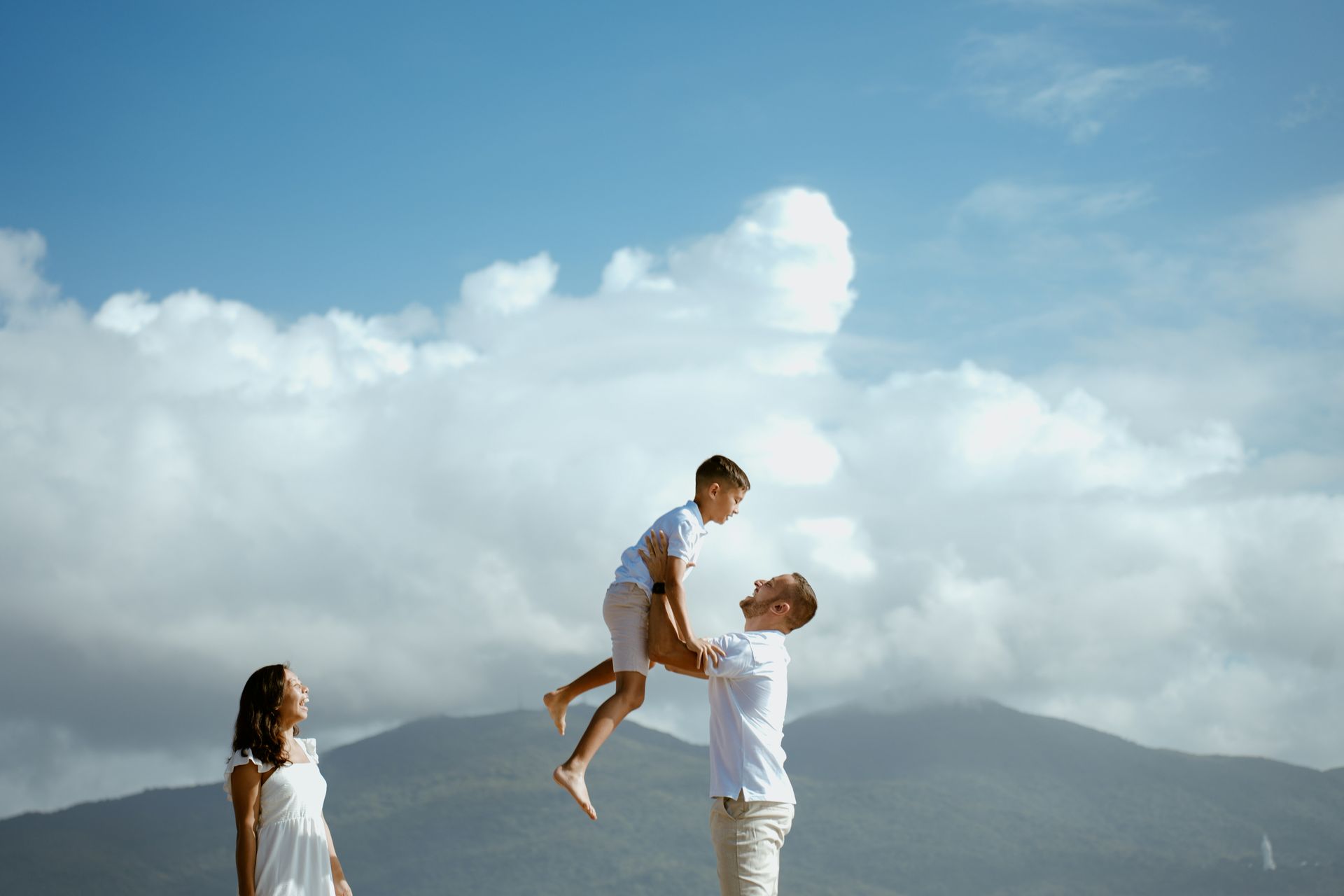 Family playing outdoors on a mountain overlook, with a man lifting a child under a cloudy blue sky.