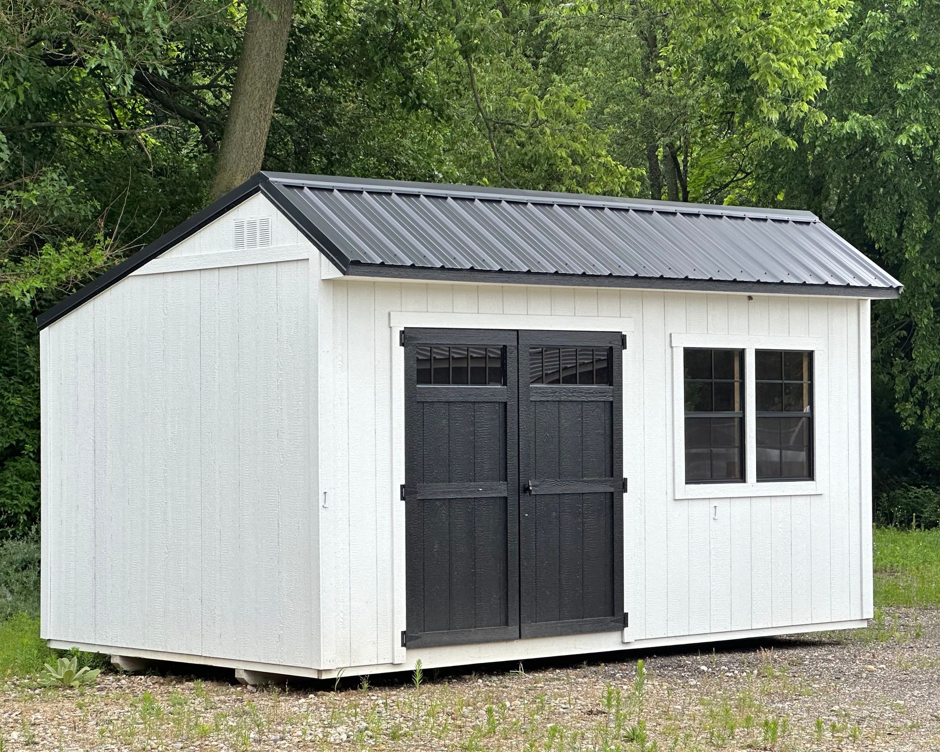 A white shed with a black roof and black doors is sitting on top of a dirt field.