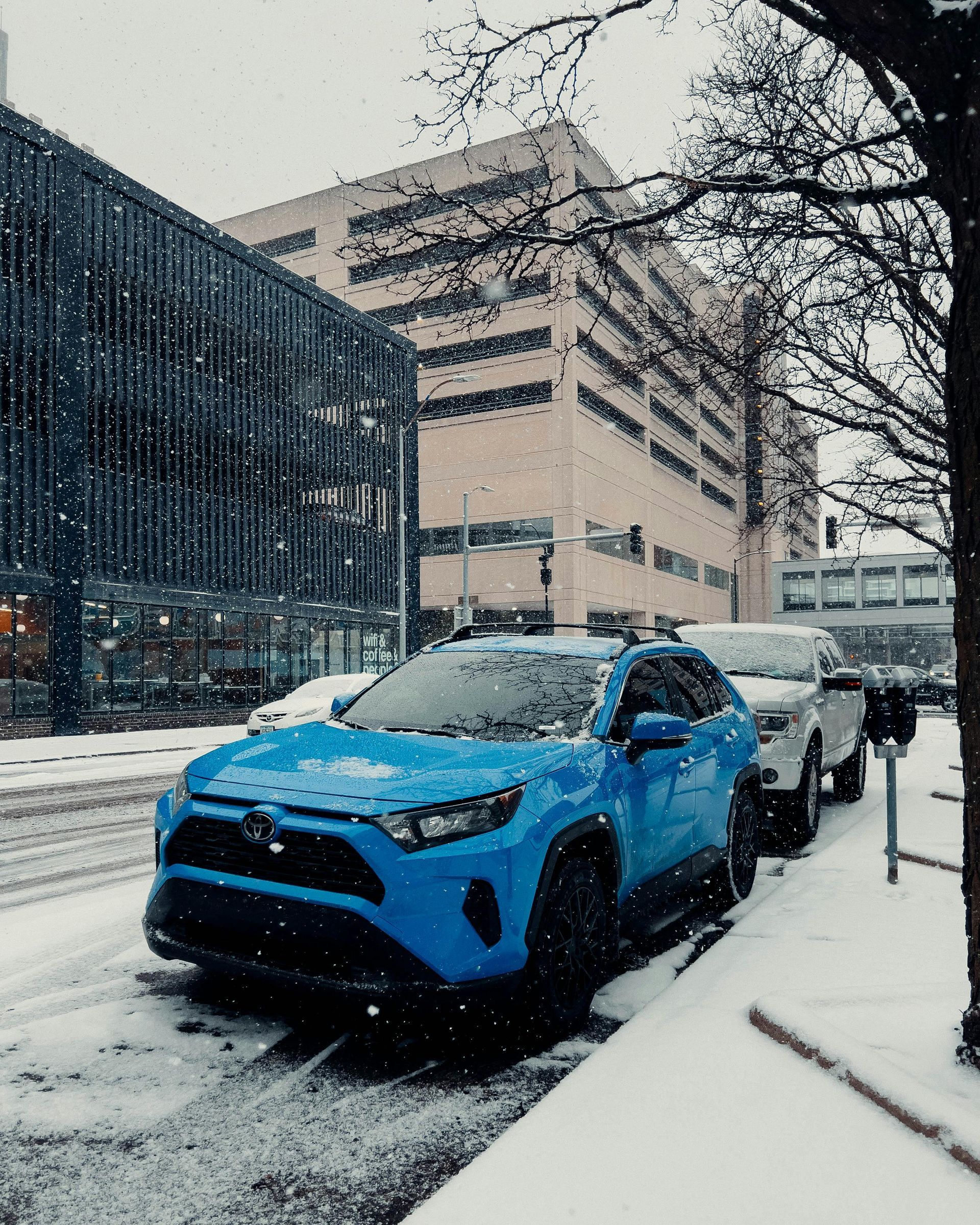 Blue SUV parked on a snowy city street beside a black car, with buildings and falling snow.