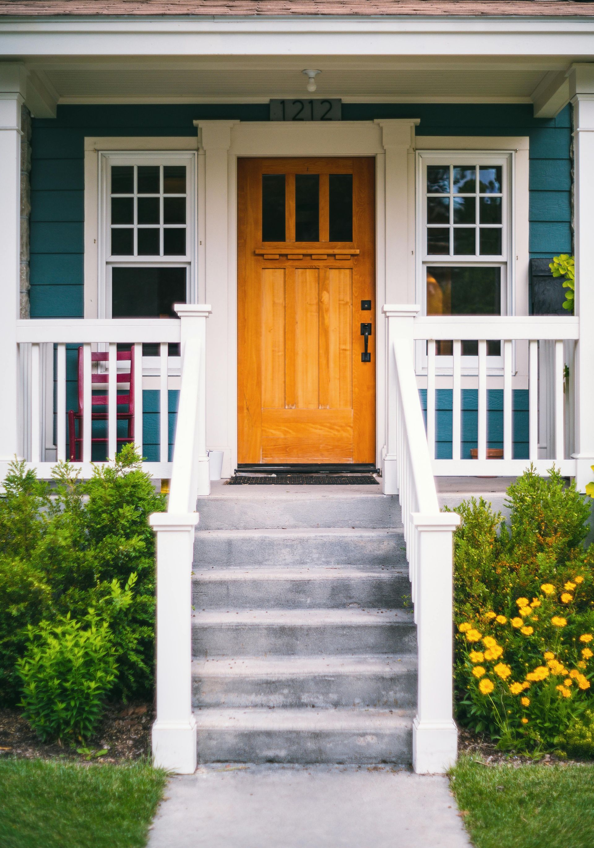 Front porch with yellow door, gray steps, white railings, and green shrubs with yellow flowers