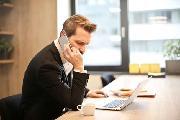 Man in a black suit talks on a phone while working on a laptop at a desk in an office.