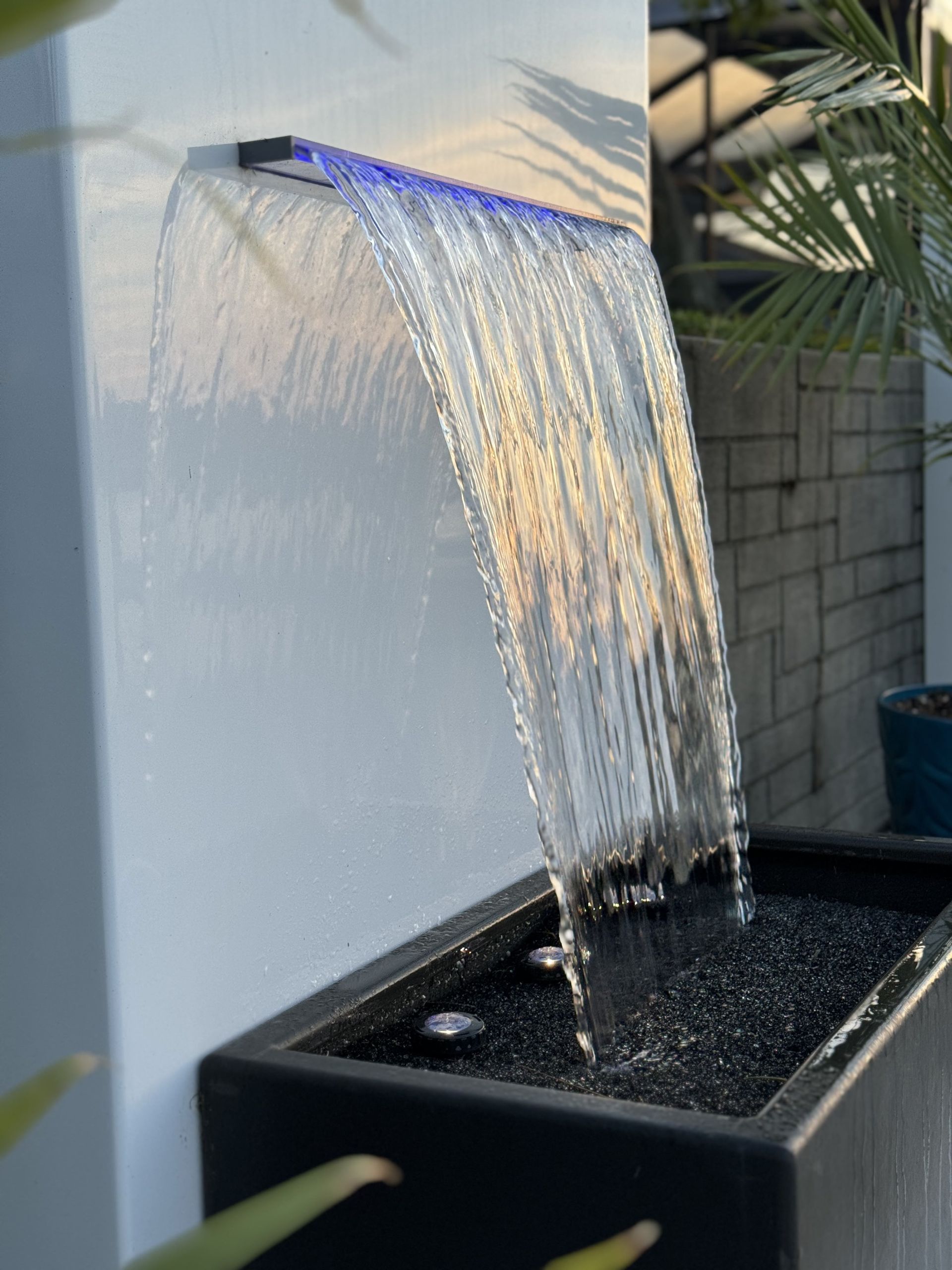 Water cascading from a wall fountain with blue LED lighting into a black basin