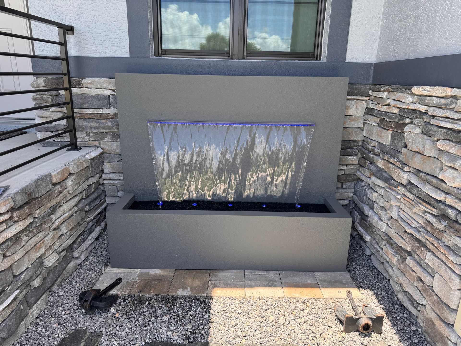 Water fountain with water cascading over a gray wall, surrounded by stone and gravel