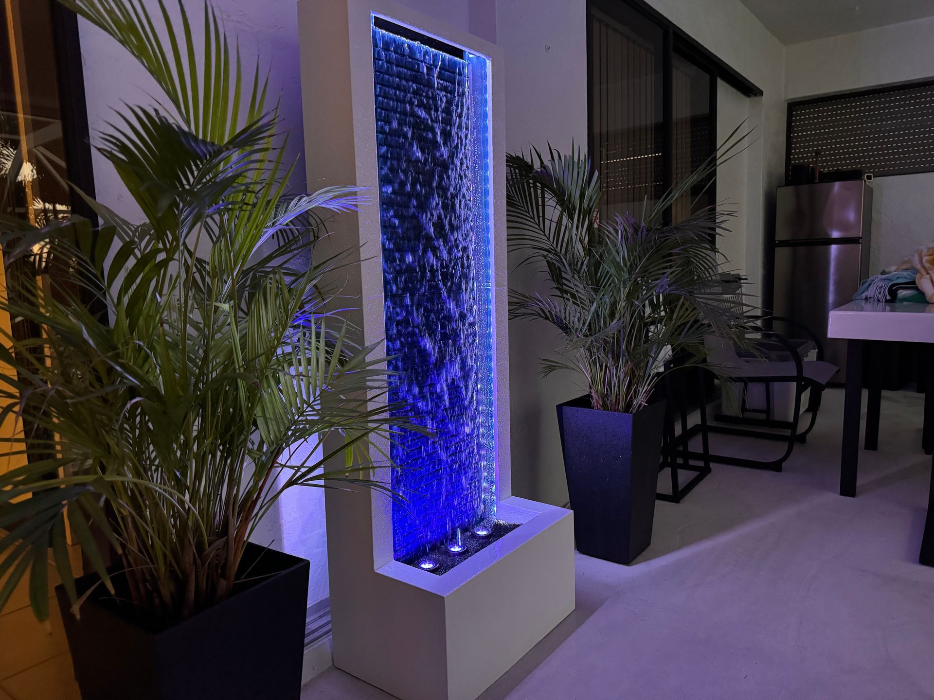 A tall water fountain with blue lighting, flanked by potted plants on a patio