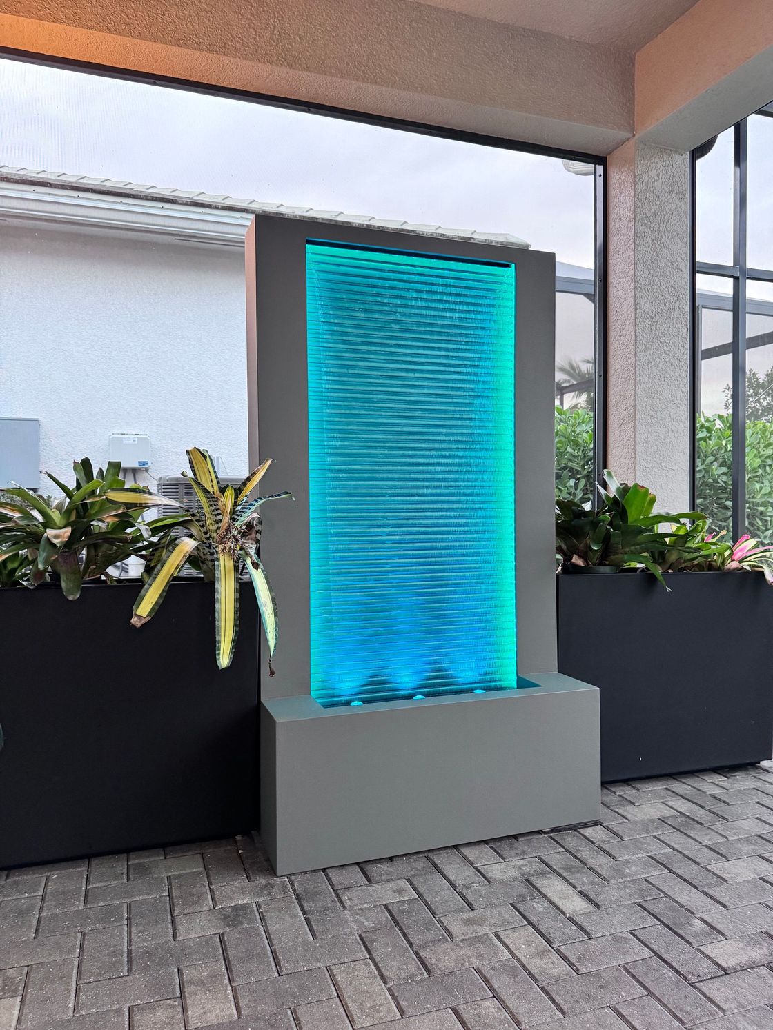 Water fountain with blue-lit rippled glass, gray frame, and planters on a brick patio