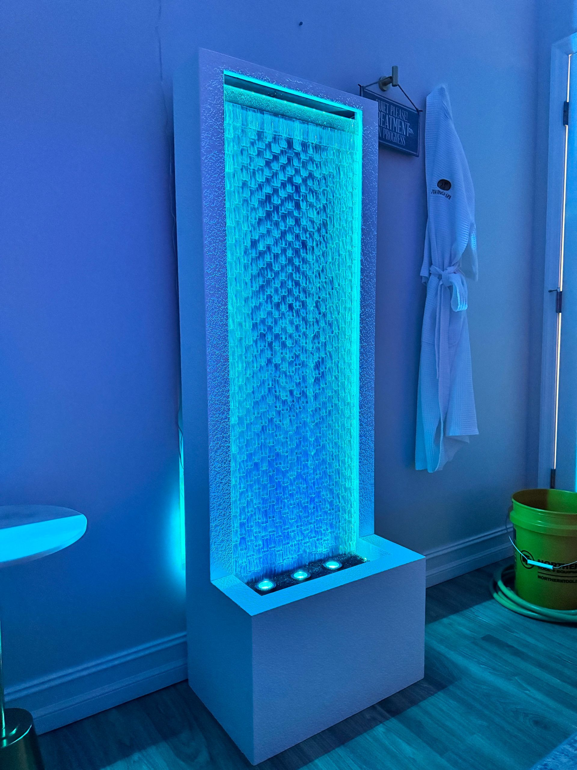 Tall indoor water fountain with blue lights, in a room with a white wall and bathrobe