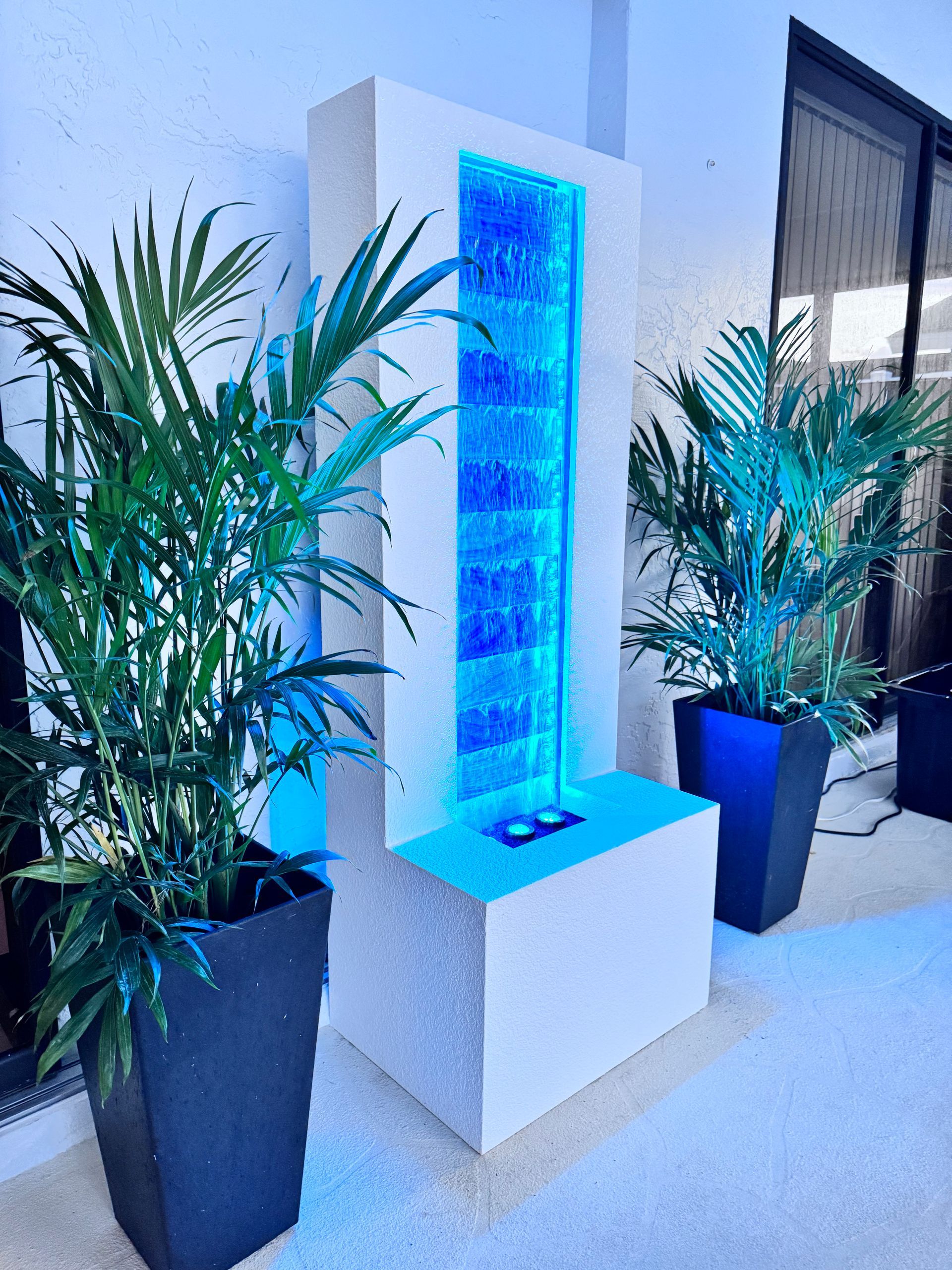 White and blue illuminated bubble water fountain with potted plants in an outdoor setting