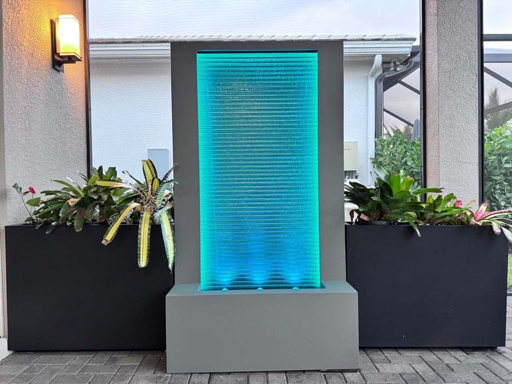 A modern outdoor water fountain with a blue-lit glass panel, flanked by dark planters with greenery.