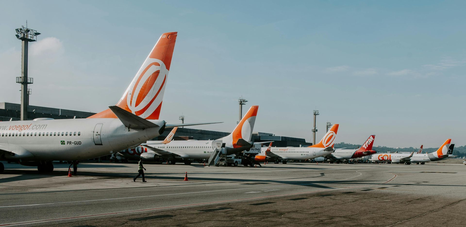 Several airplanes parked on the tarmac. The planes have orange and white designs. A person walks in front of the planes.
