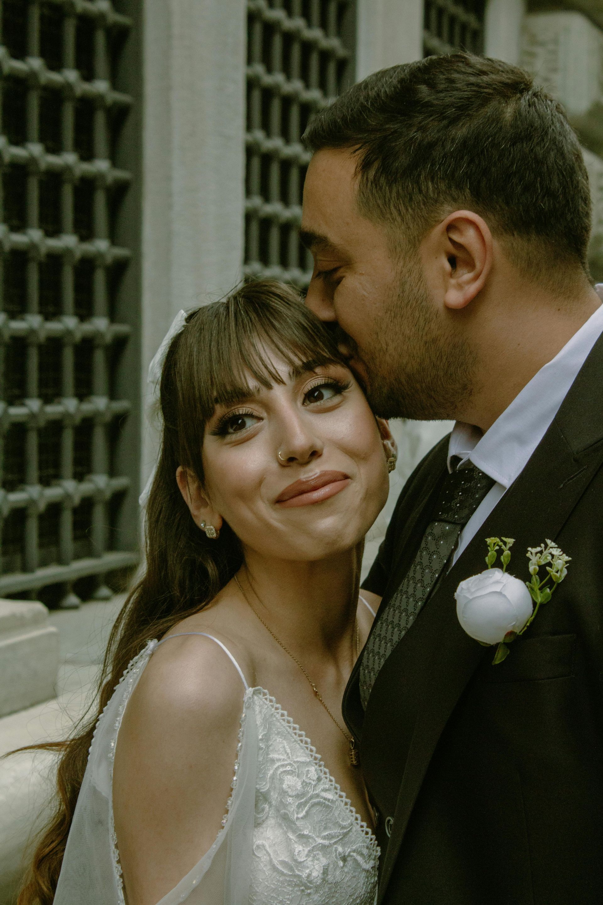 Groom kisses bride's forehead. They are outdoors, near a building with a window. The bride wears a white dress and veil.