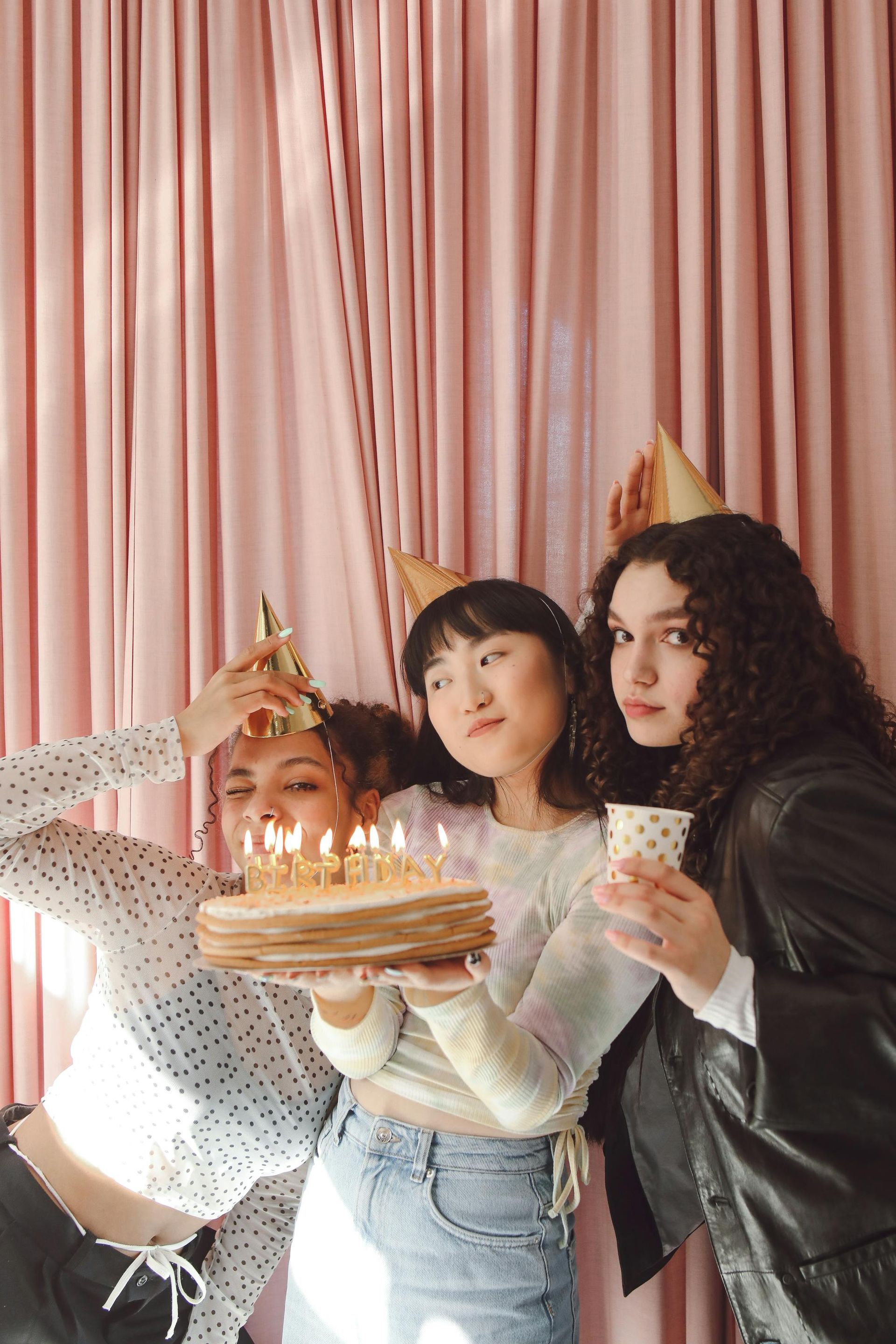 Three people celebrating with a birthday cake; pink curtains in background.