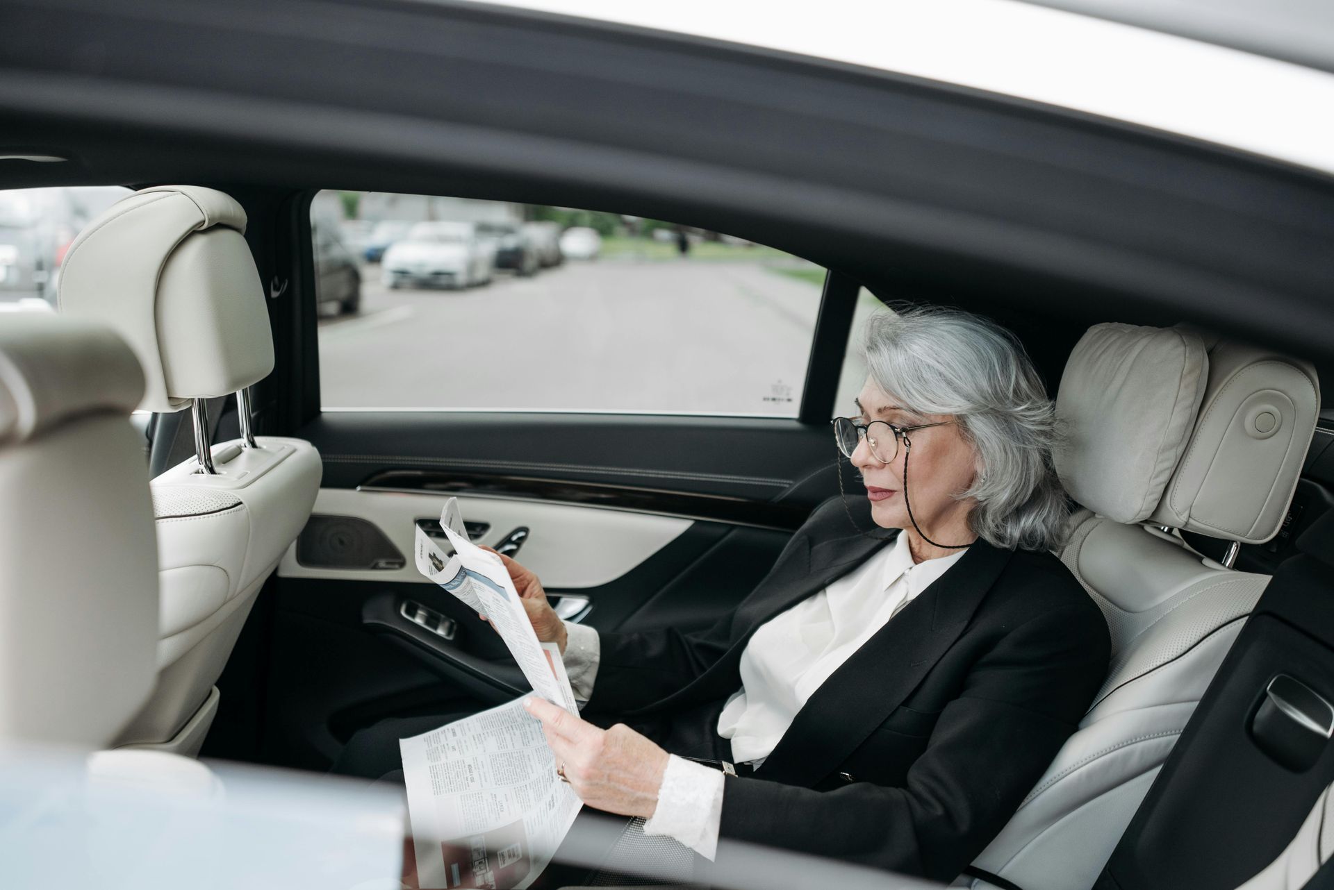 Woman with gray hair wearing glasses reads papers in the back seat of a car.