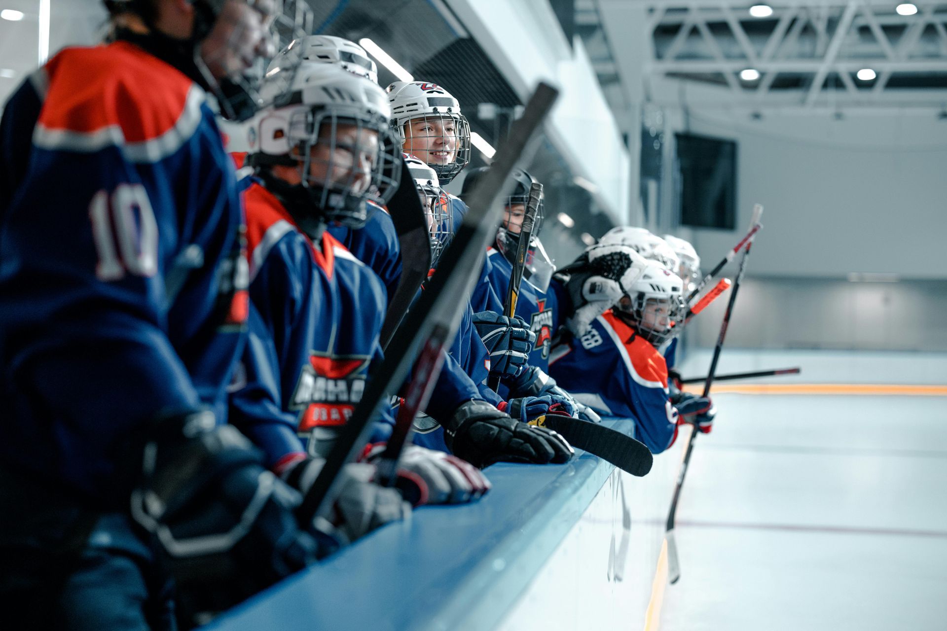 Hockey players in blue and red jerseys on a rink's bench, helmets on, holding hockey sticks.