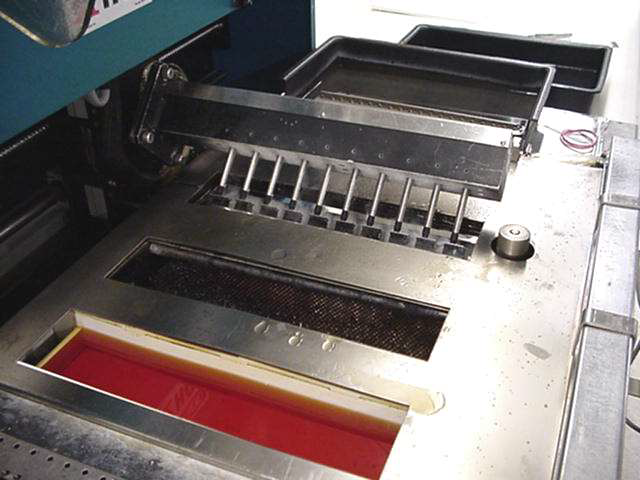 A lab machine with a row of nozzles over a rectangular tray filled with red liquid, set on a stainless steel surface.