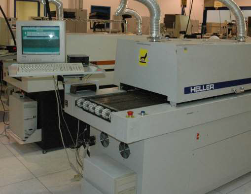 A Heller reflow soldering machine with a conveyor belt and connected computer terminal in a manufacturing facility.