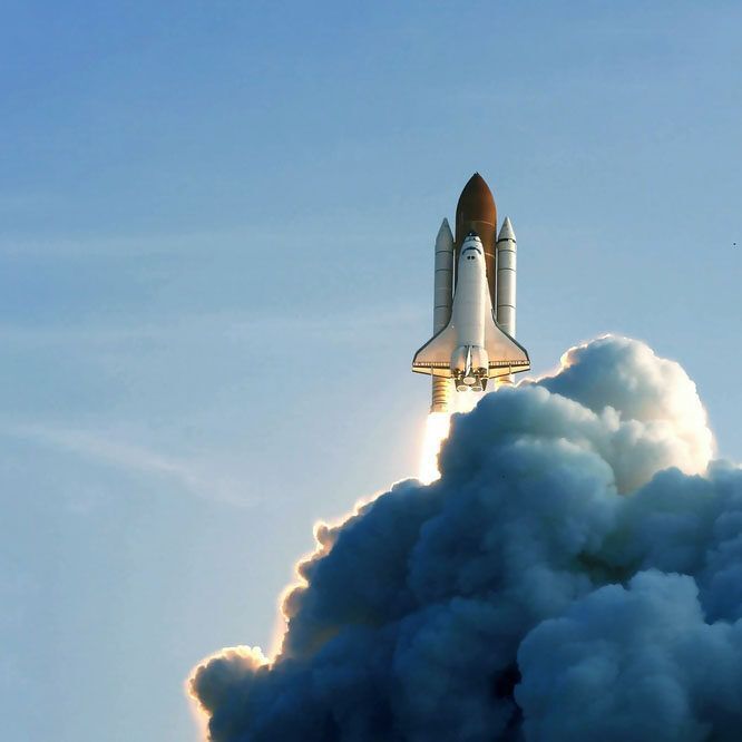Space shuttle launching, ascending through white smoke, against a blue sky.