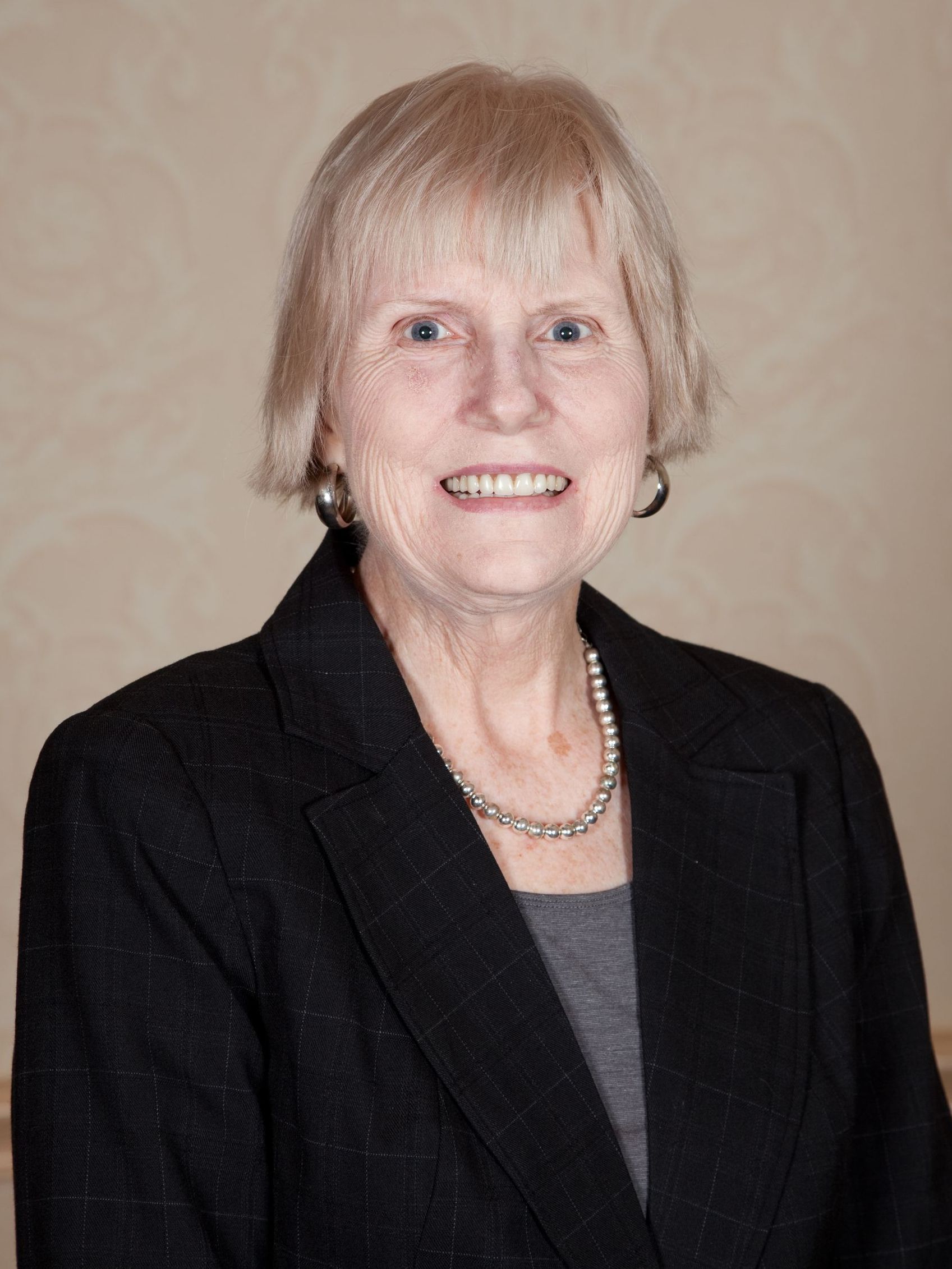 A woman wearing a black jacket and a pearl necklace is smiling for the camera.