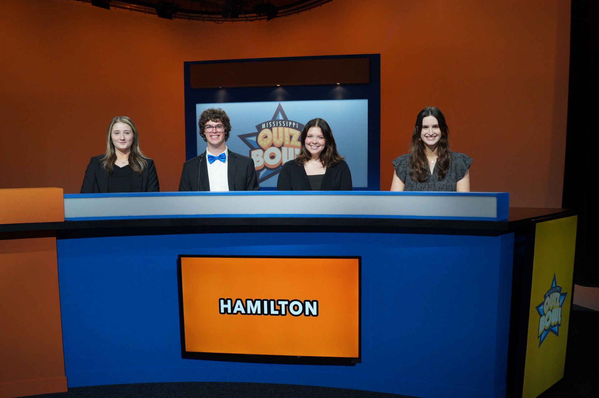 A group of people are sitting at a desk in front of a screen that says hamilton.