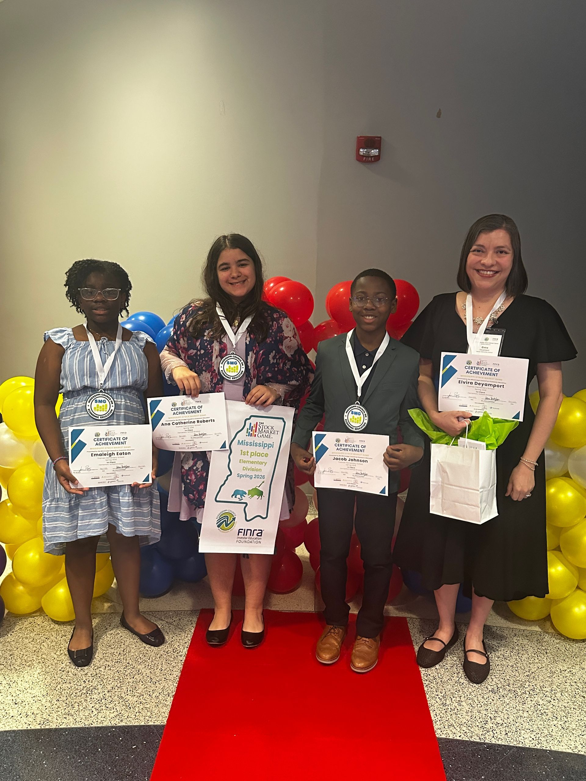 Four people holding certificates on a red carpet, posing in front of red, blue and yellow balloons.