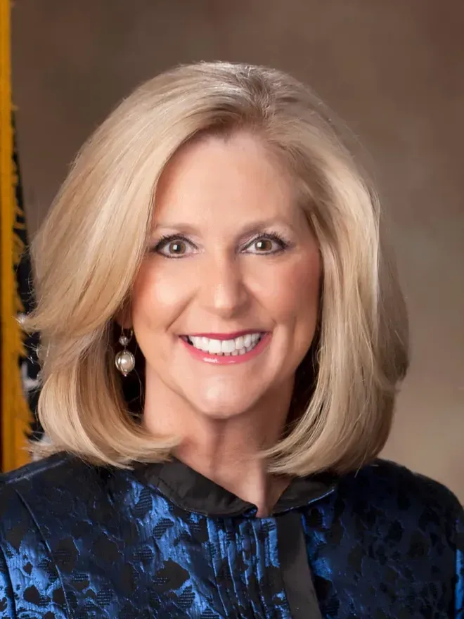 A woman in a blue jacket is smiling in front of a flag.