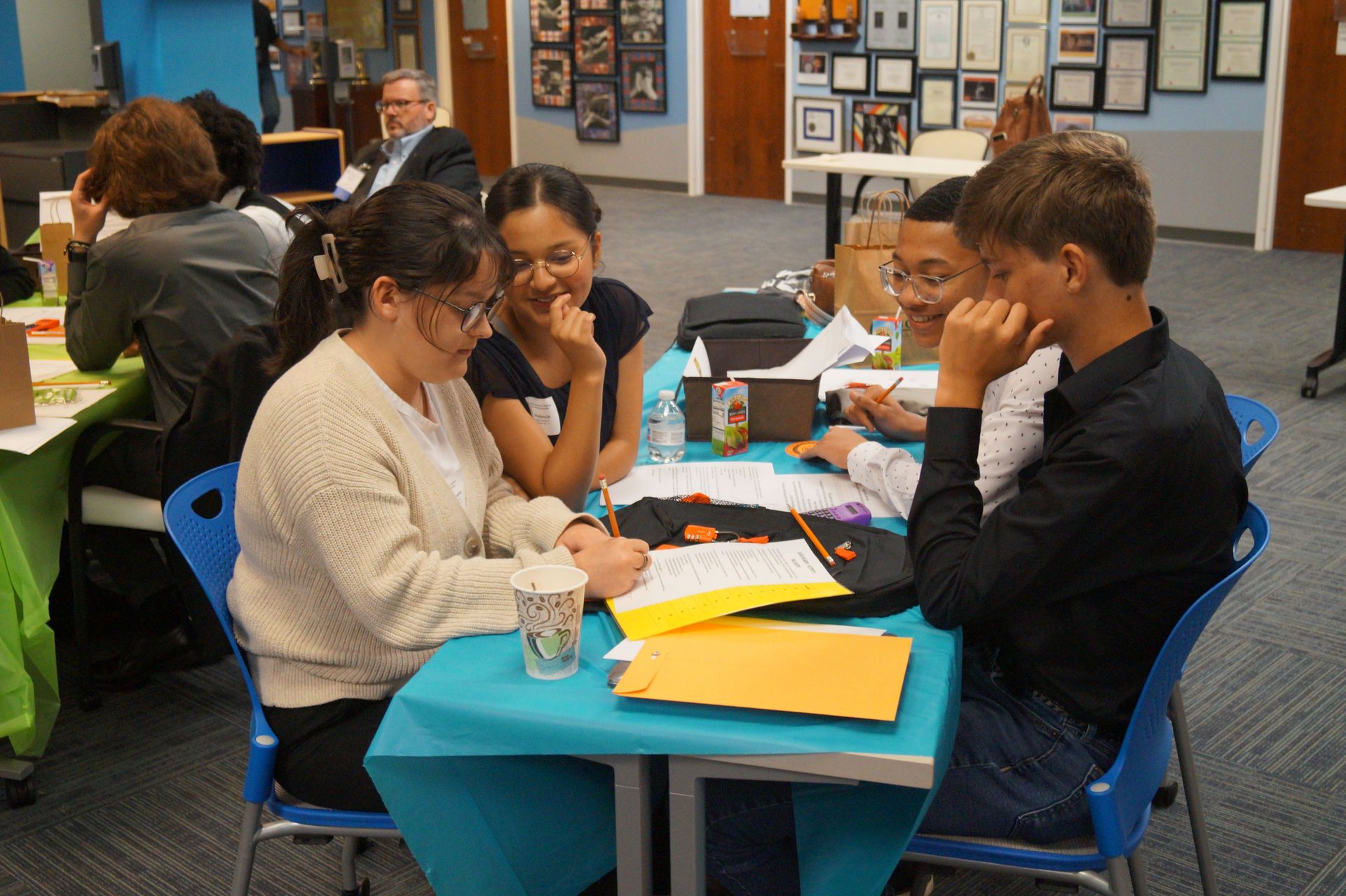 Group of people at a table, looking at papers. Some smiling, focused, and sitting in blue chairs, indoor setting.
