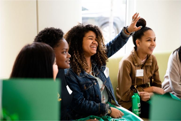 A group of young people are sitting around a table with one girl raising her hand.