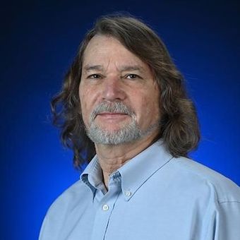 A man with long hair and a beard is standing in front of a blue background.