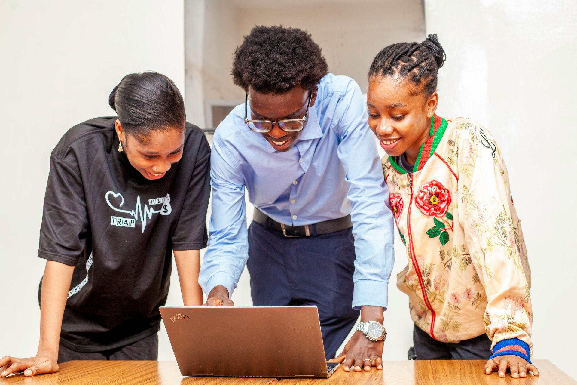 Three people lean over a laptop on a table, looking at the screen together.