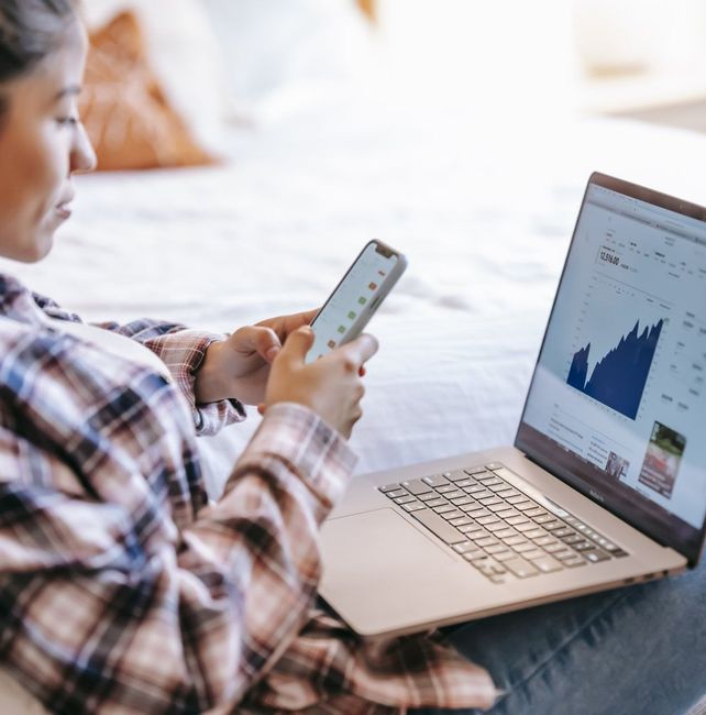 A woman is sitting in front of a laptop and looking at her phone