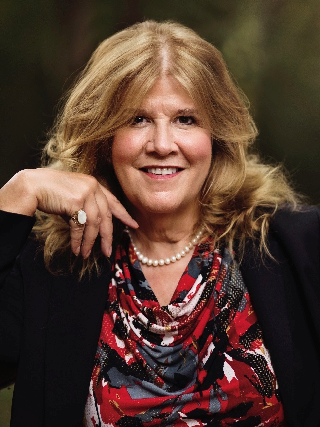 A woman wearing a pearl necklace and a black jacket is smiling.