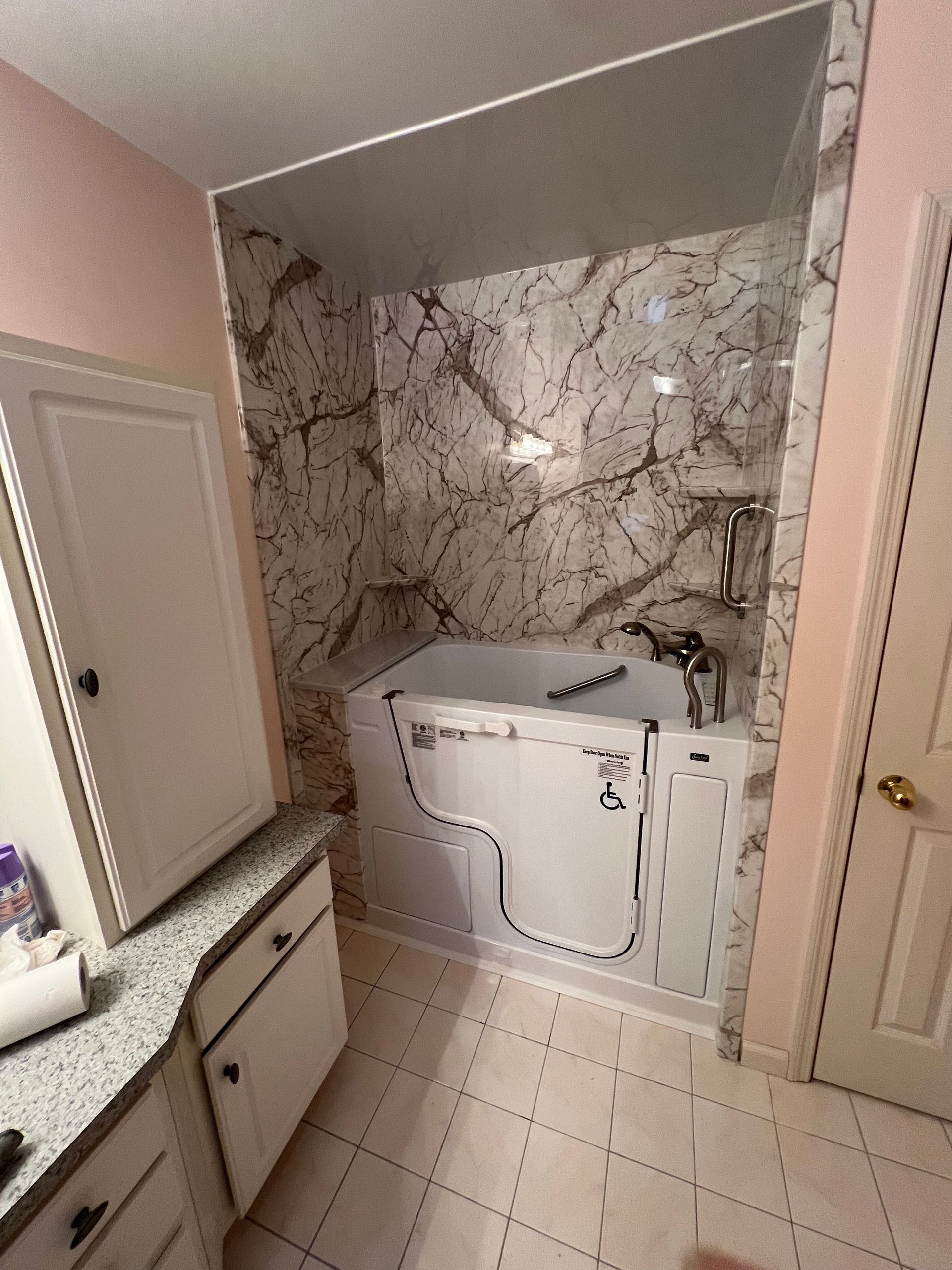 A white walk-in bathtub installed in a marble-tiled alcove in a bathroom with a white vanity and tiled flooring.