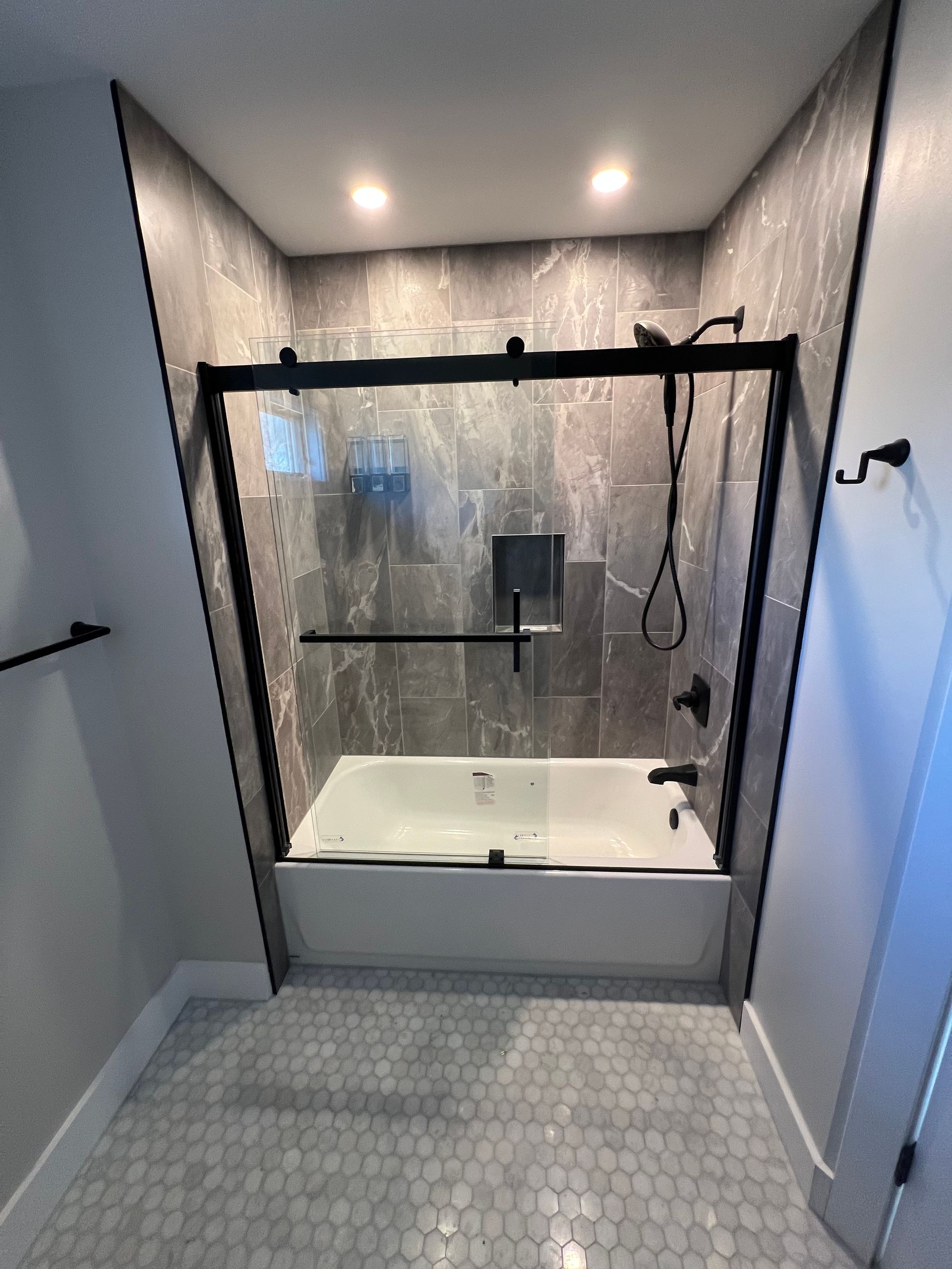 Modern bathroom with a bathtub and shower enclosure. Gray tile walls, black fixtures, and gray hexagon floor tiles.