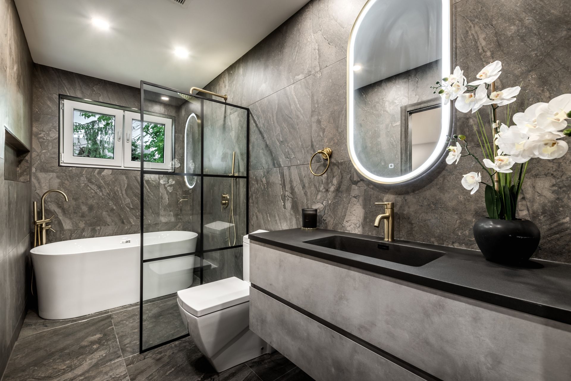Modern bathroom with gray textured walls, gold fixtures, black countertop, glass shower, and freestanding white bathtub.