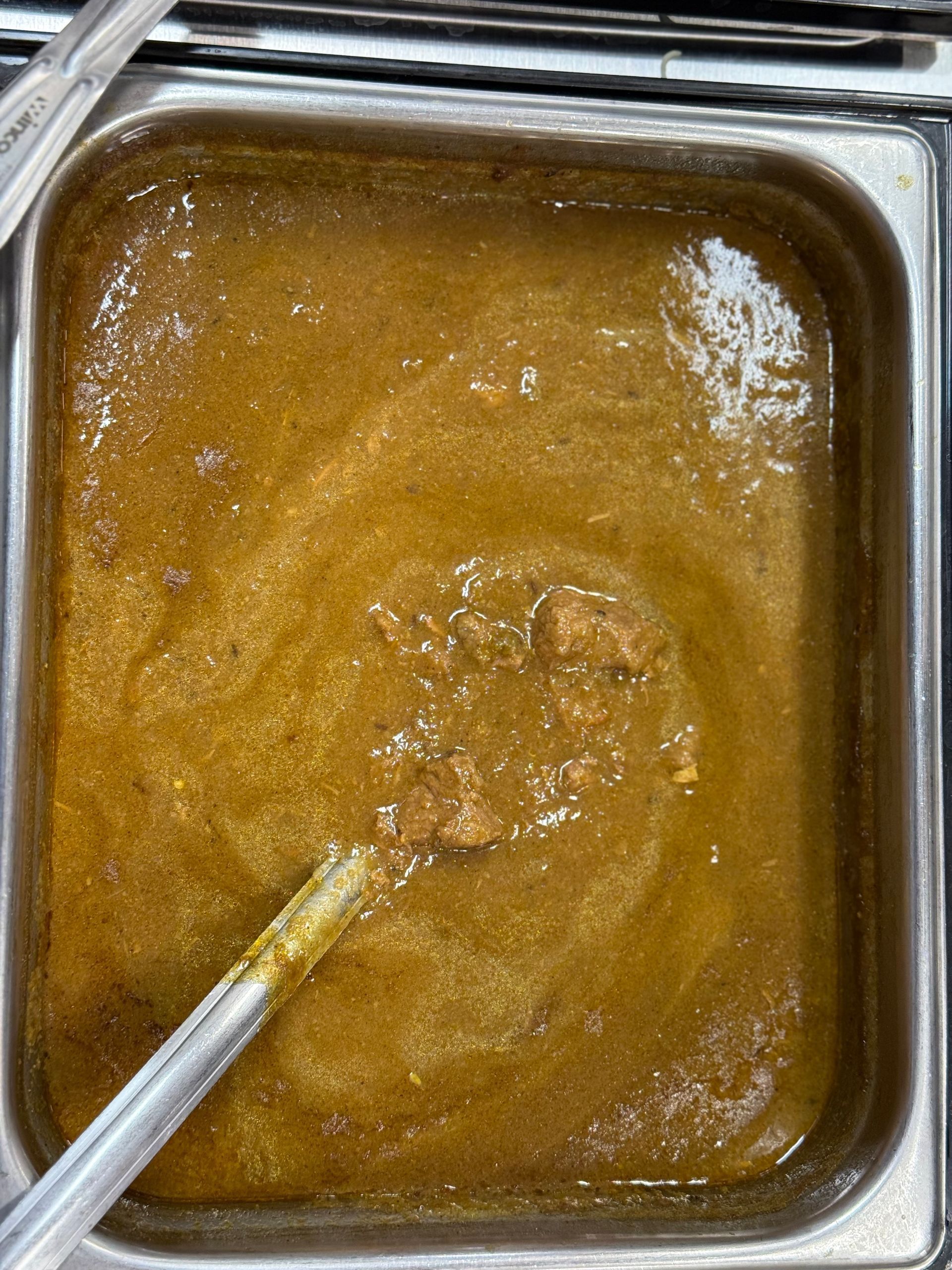 Stew in a metal pan with a serving spoon. Brown liquid with visible chunks.