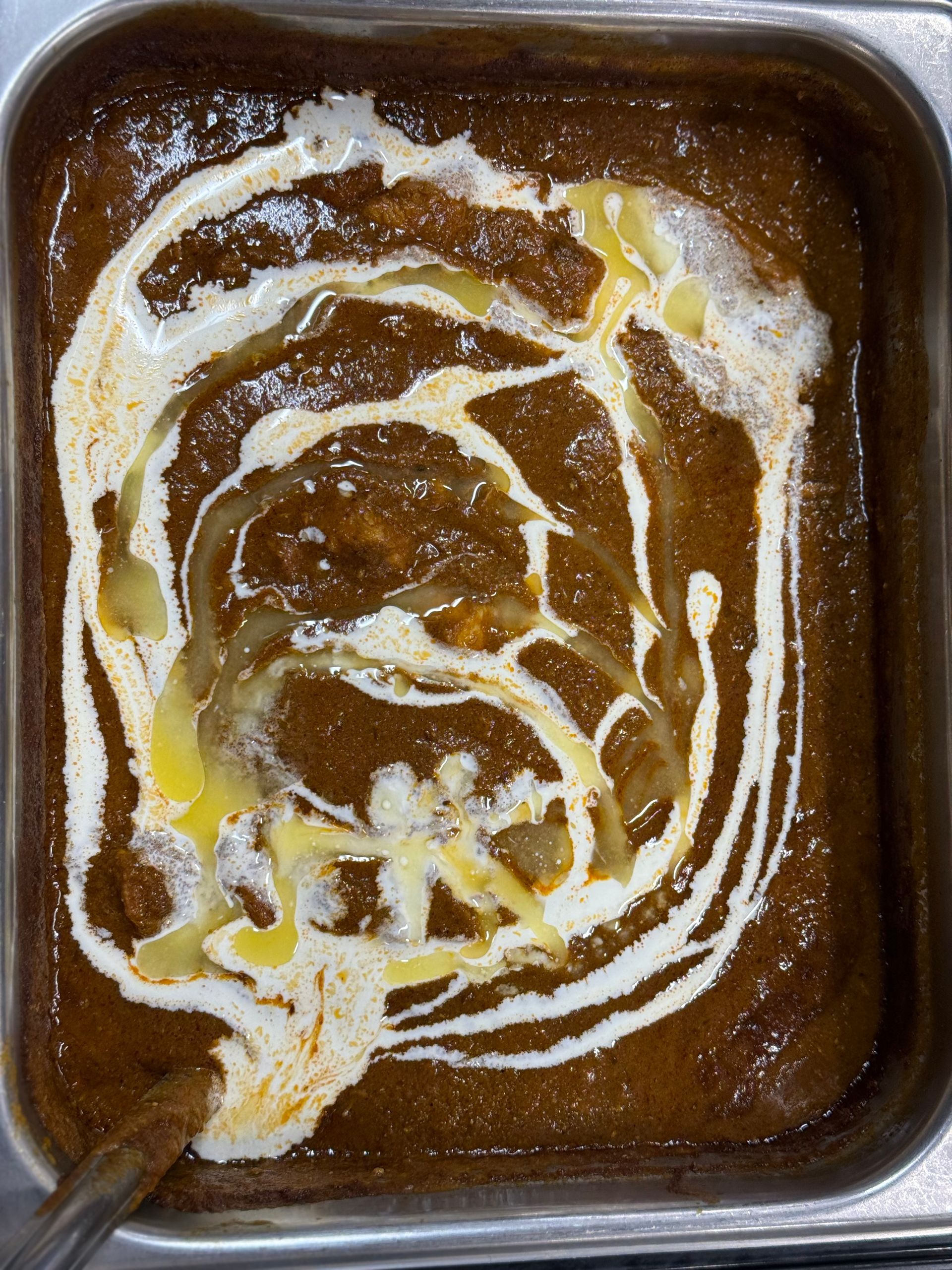 Dark brown curry topped with swirls of white cream and yellow butter in a metal container.