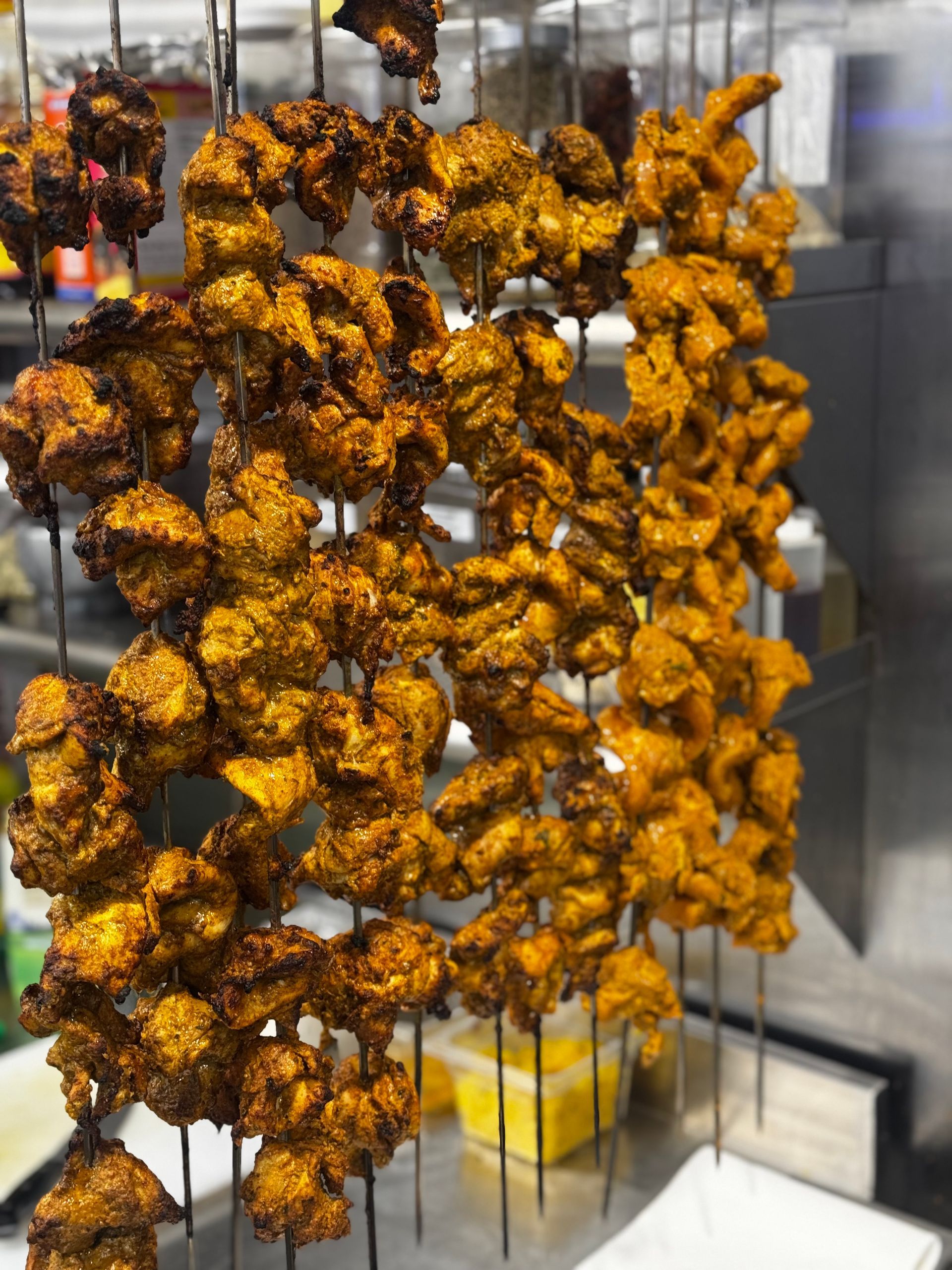 Skewered, seasoned chicken pieces, likely tandoori, hanging in a commercial kitchen. Golden-brown color.
