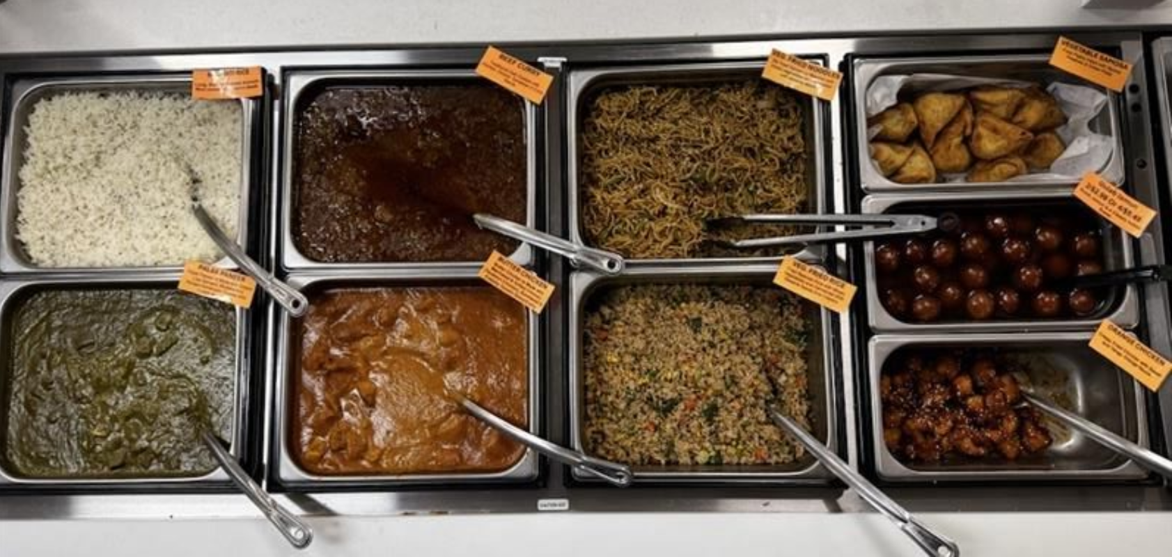 Food buffet with eight different dishes, each in a metal tray, with serving utensils and labels.