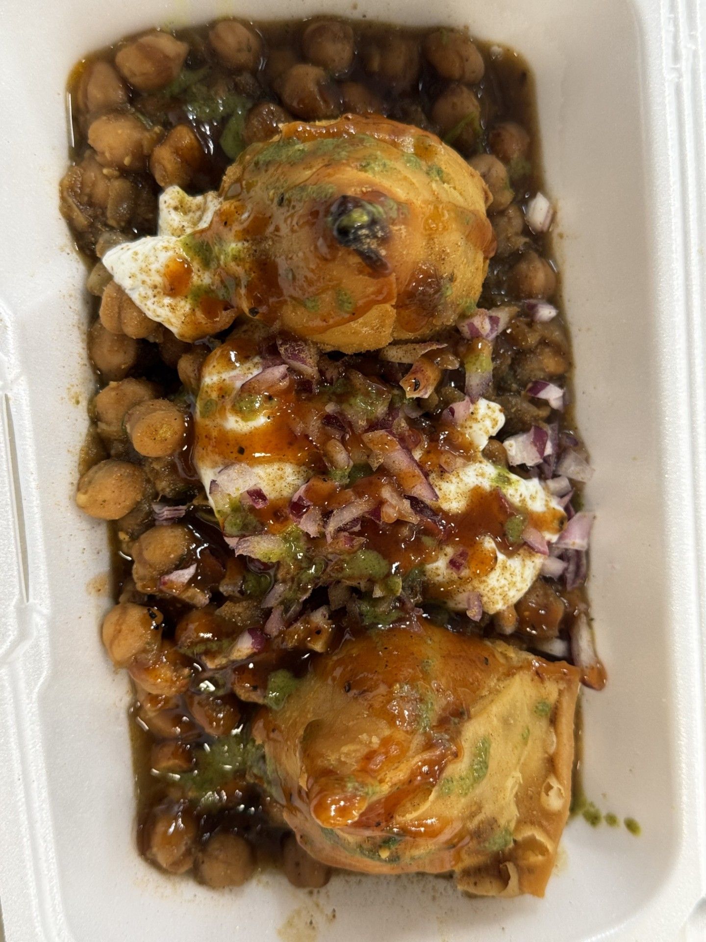 Chickpea chaat with fried pastries, yogurt, onions, and sauces in a white takeout container.