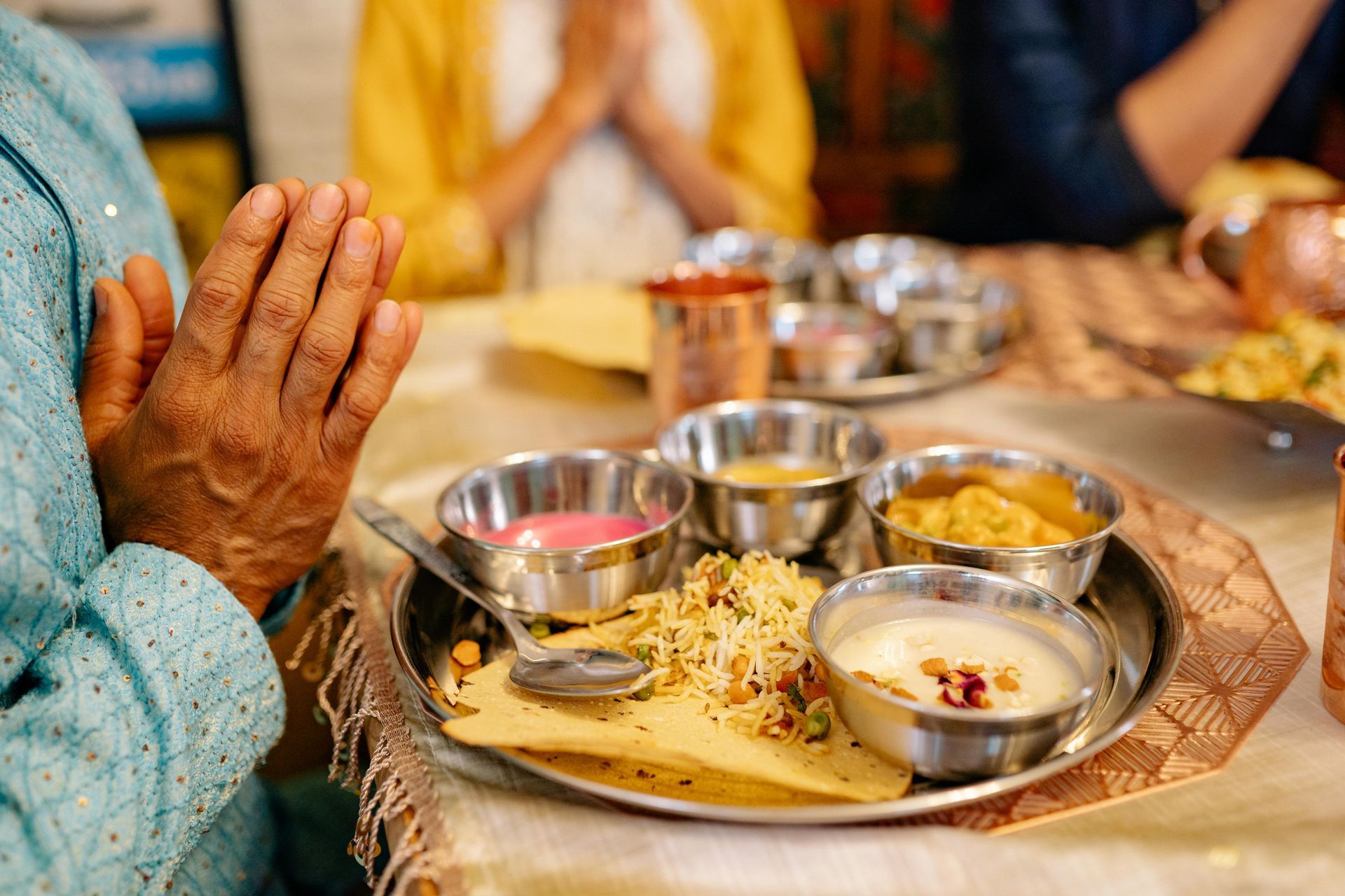 Person with clasped hands over a plate of Indian food, other people out of focus.