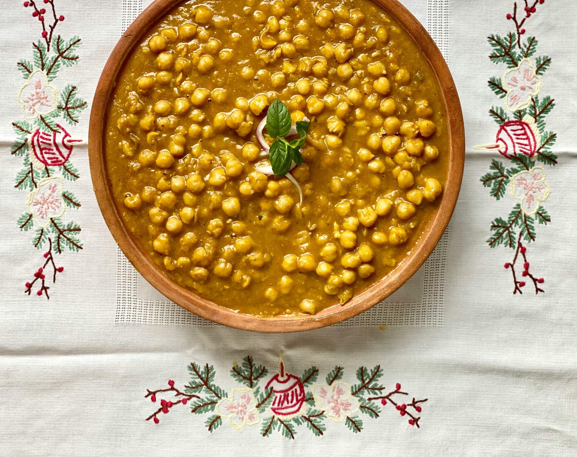 Bowl of chickpea curry, golden-brown, garnished with mint, on a festive embroidered tablecloth.