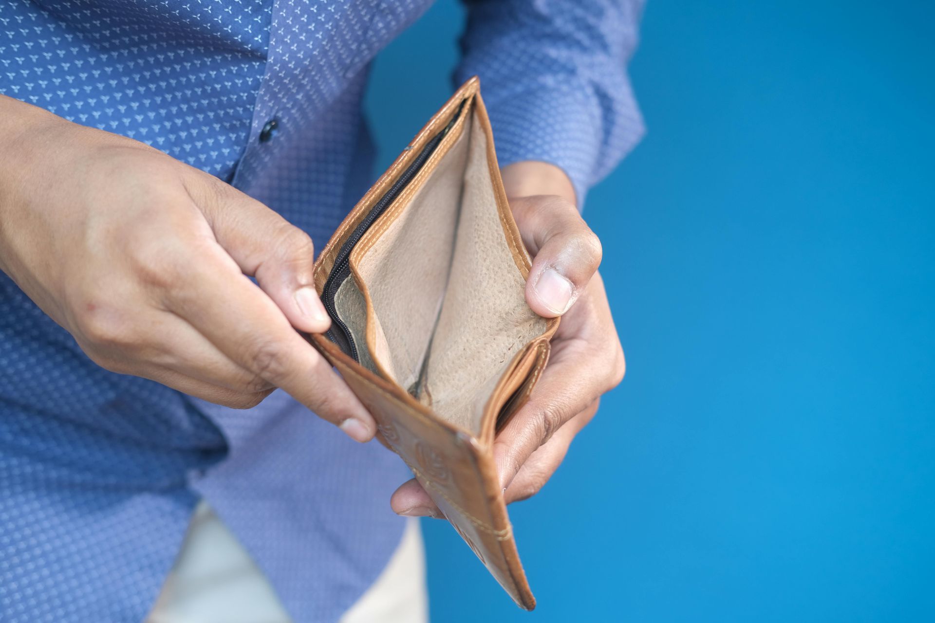 Person holding an empty brown wallet against a blue background, showing financial hardship.