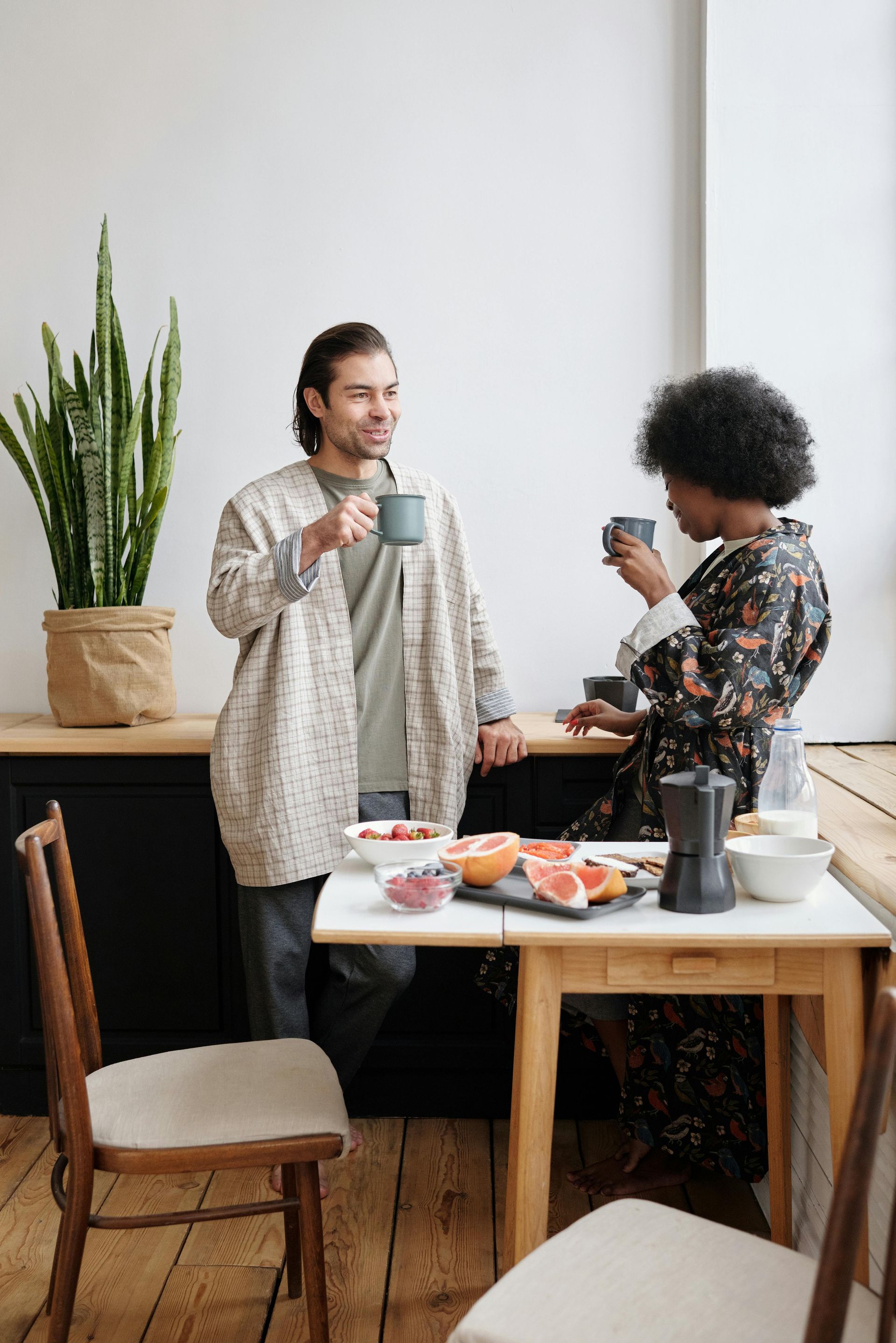 A couple drinks coffee by a table with fruit, a plant, and a coffee maker in a kitchen setting.