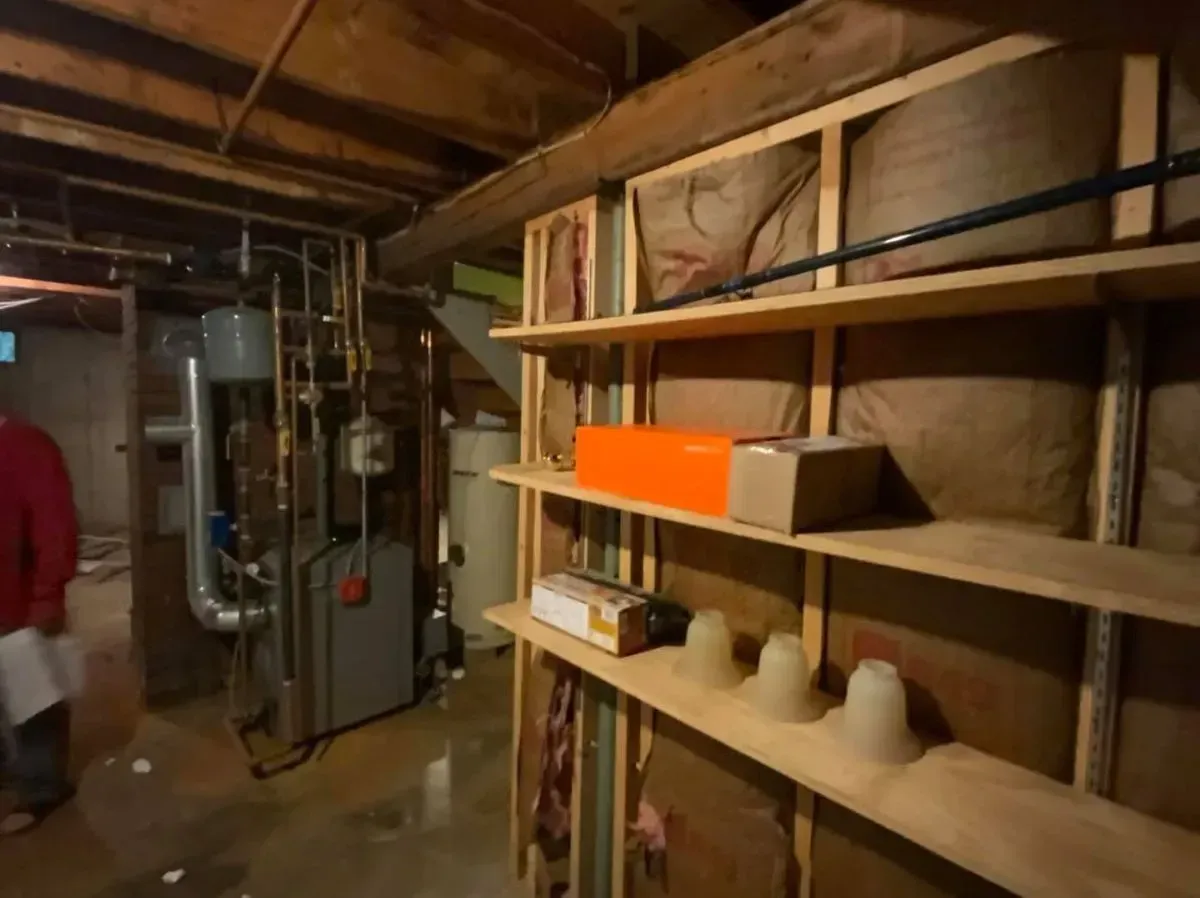 A man in a red shirt is standing in a basement with shelves and a heater.