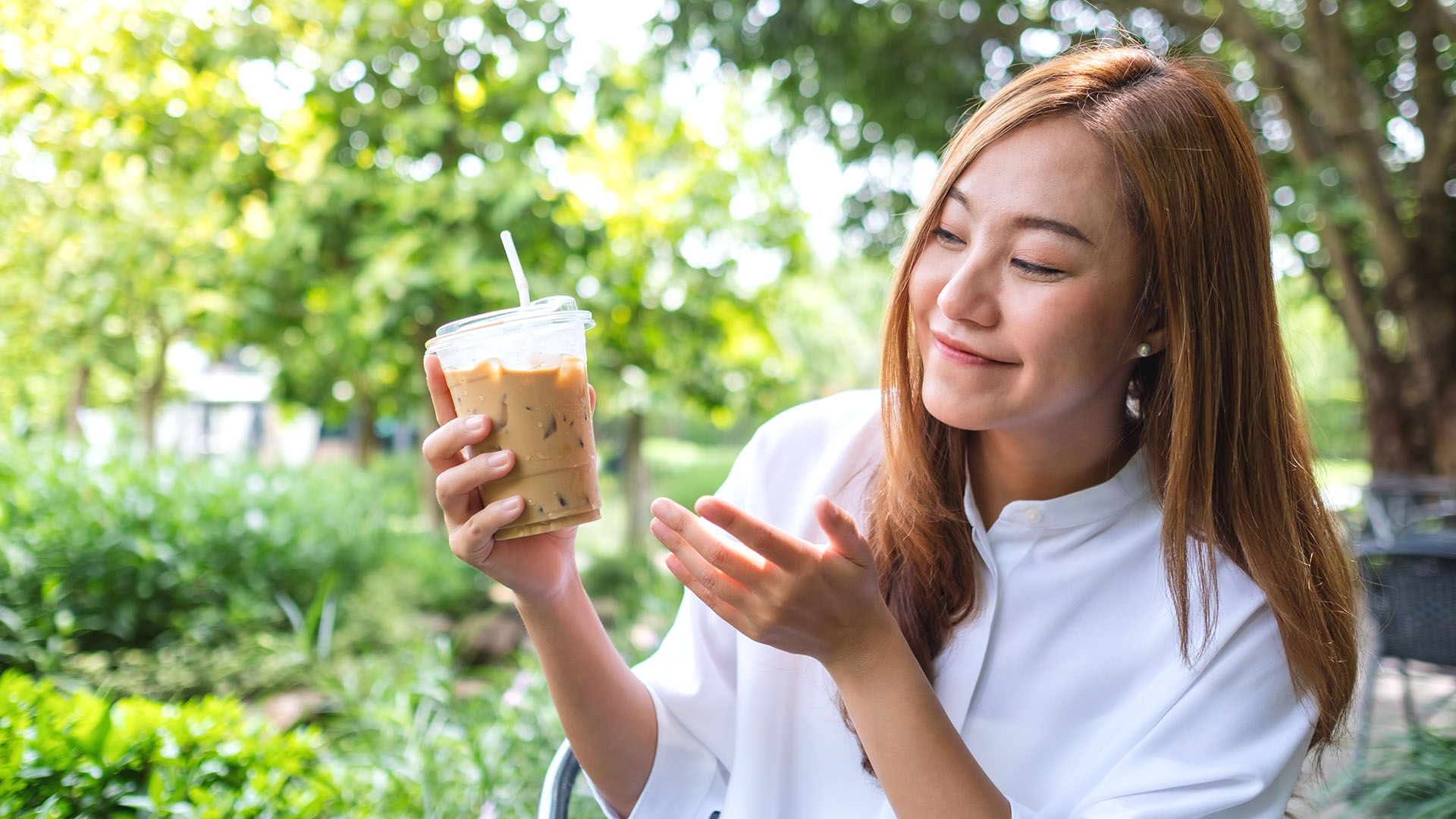 Woman smiling, holding an iced coffee drink with a straw, outdoors near greenery.