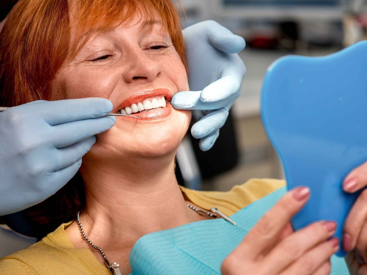 A woman is sitting in a dental chair looking at her teeth in a mirror.